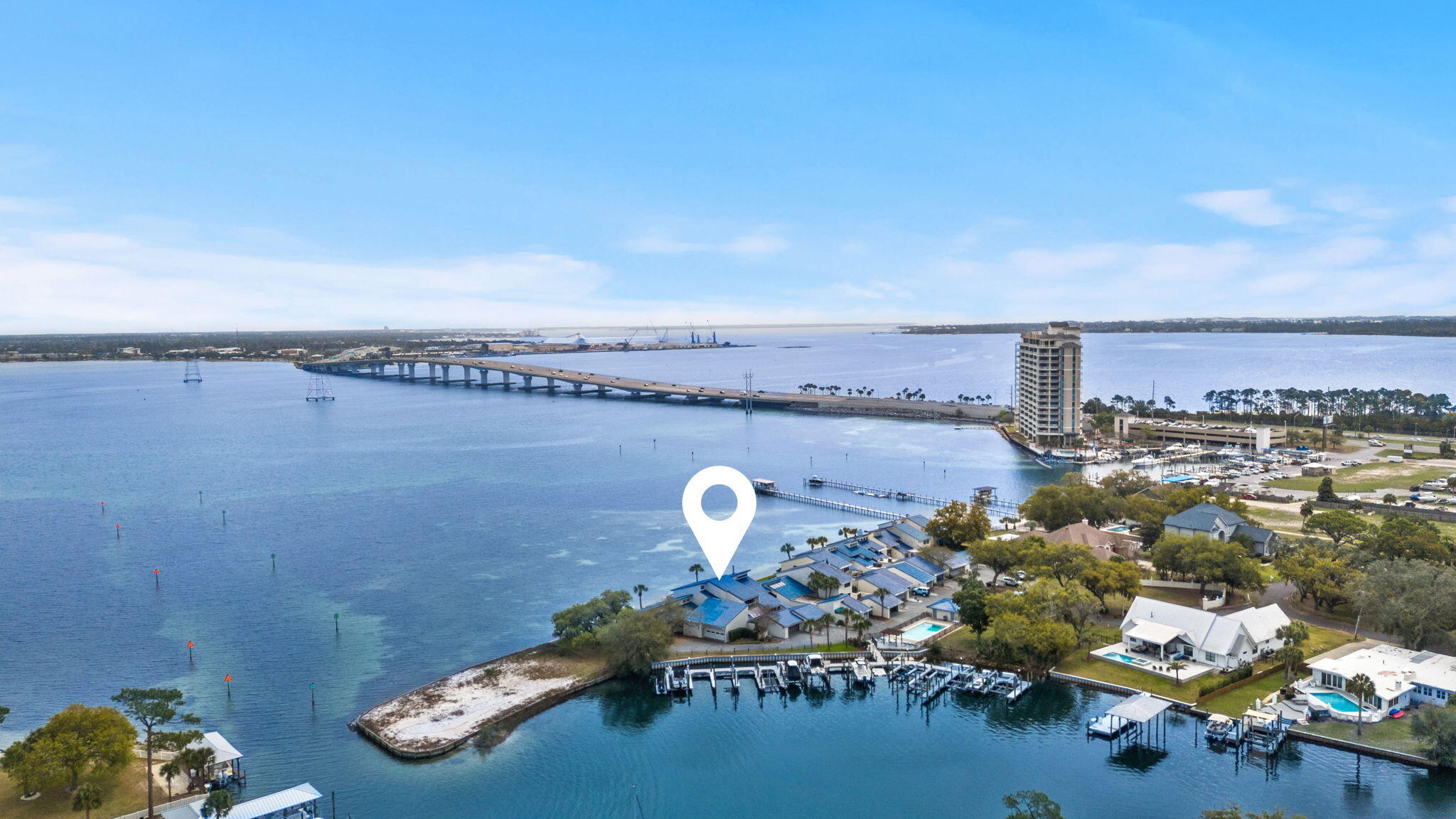 Aerial view of waterfront homes marked by a white pin, with marinas and a long bridge across the bay.