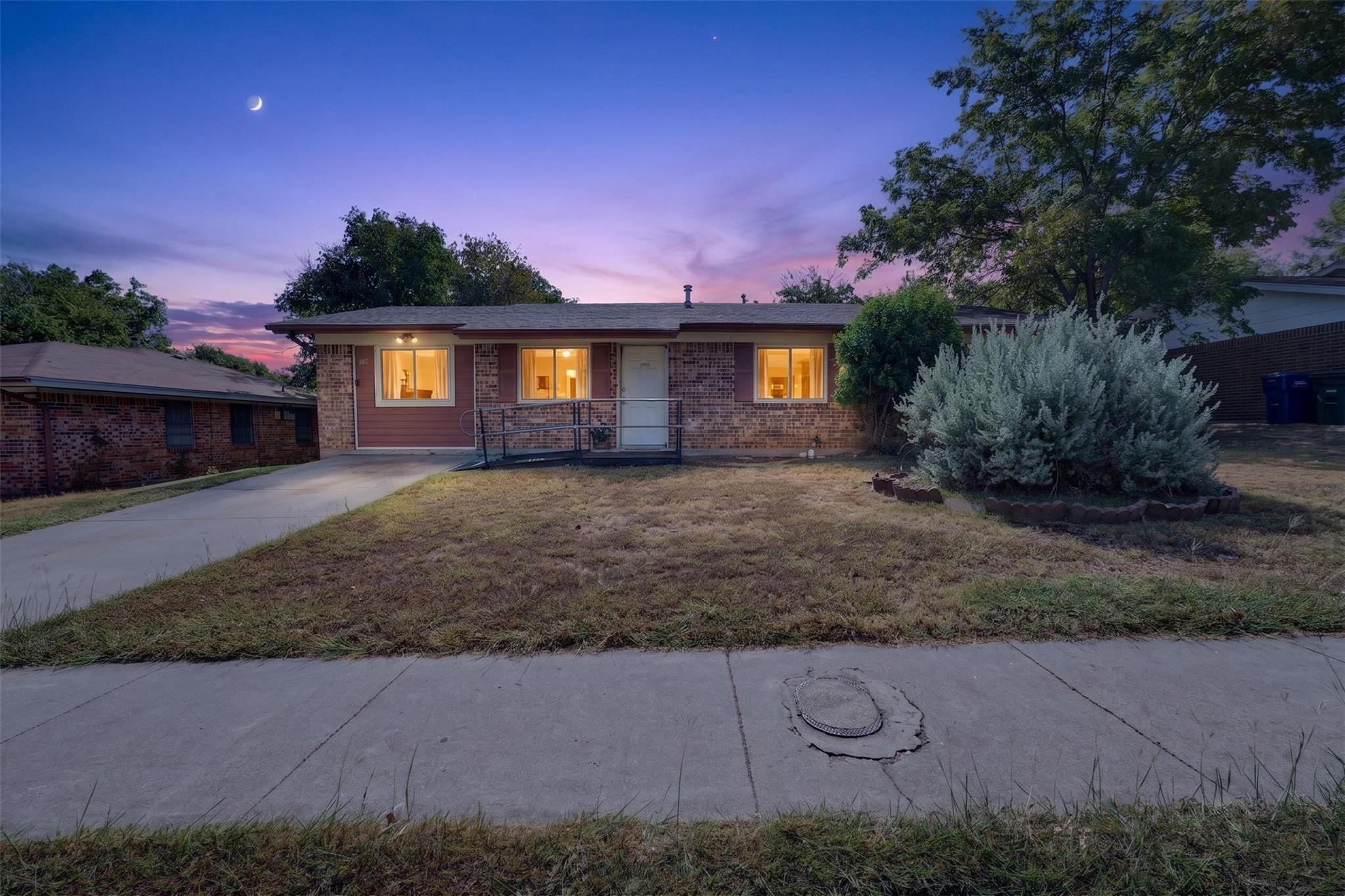 Single-story brick house at dusk with glowing windows, an access ramp, and a large front-yard shrub.