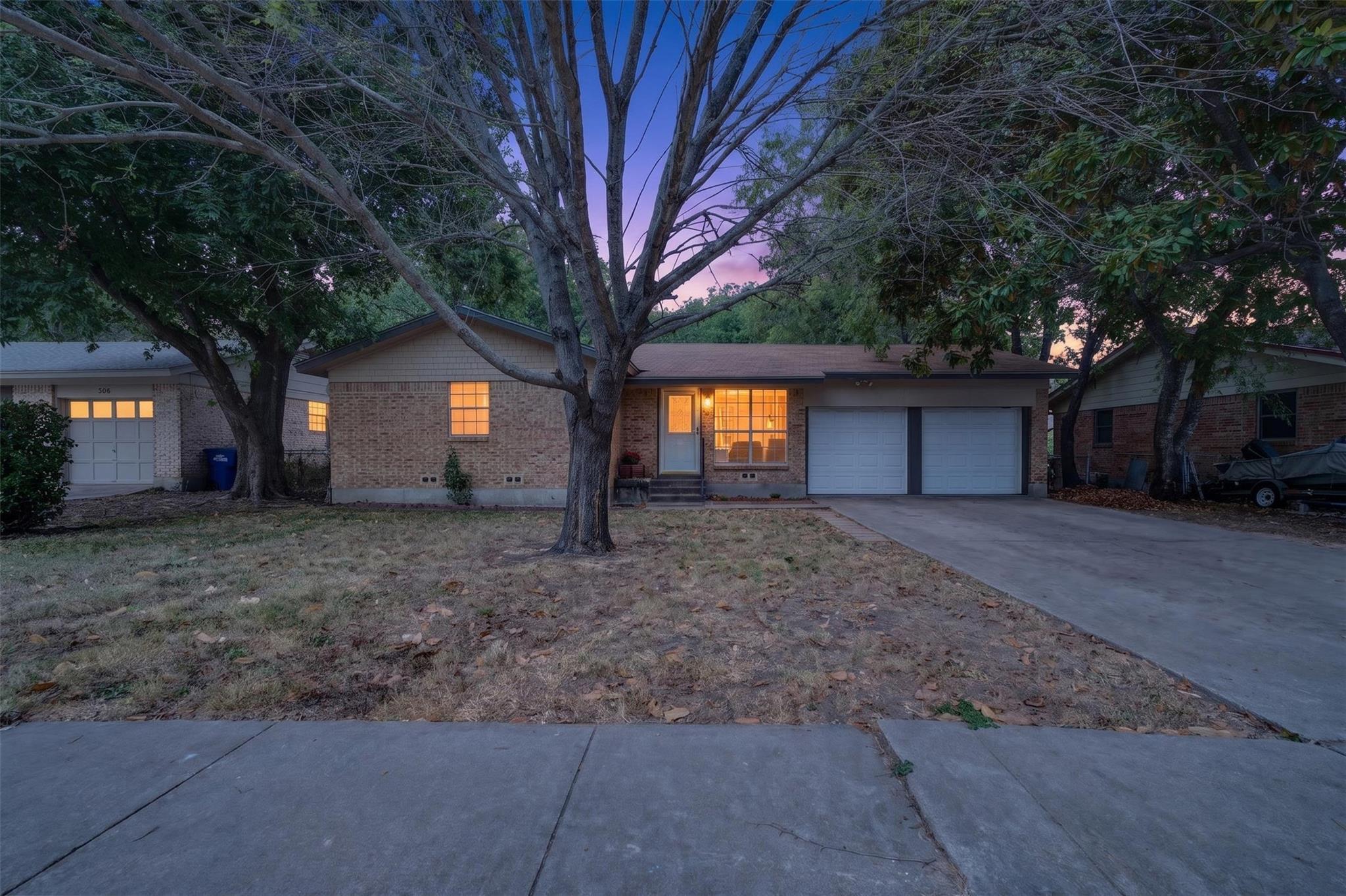 Brick house at dusk with glowing windows, a two-car garage, and mature trees.