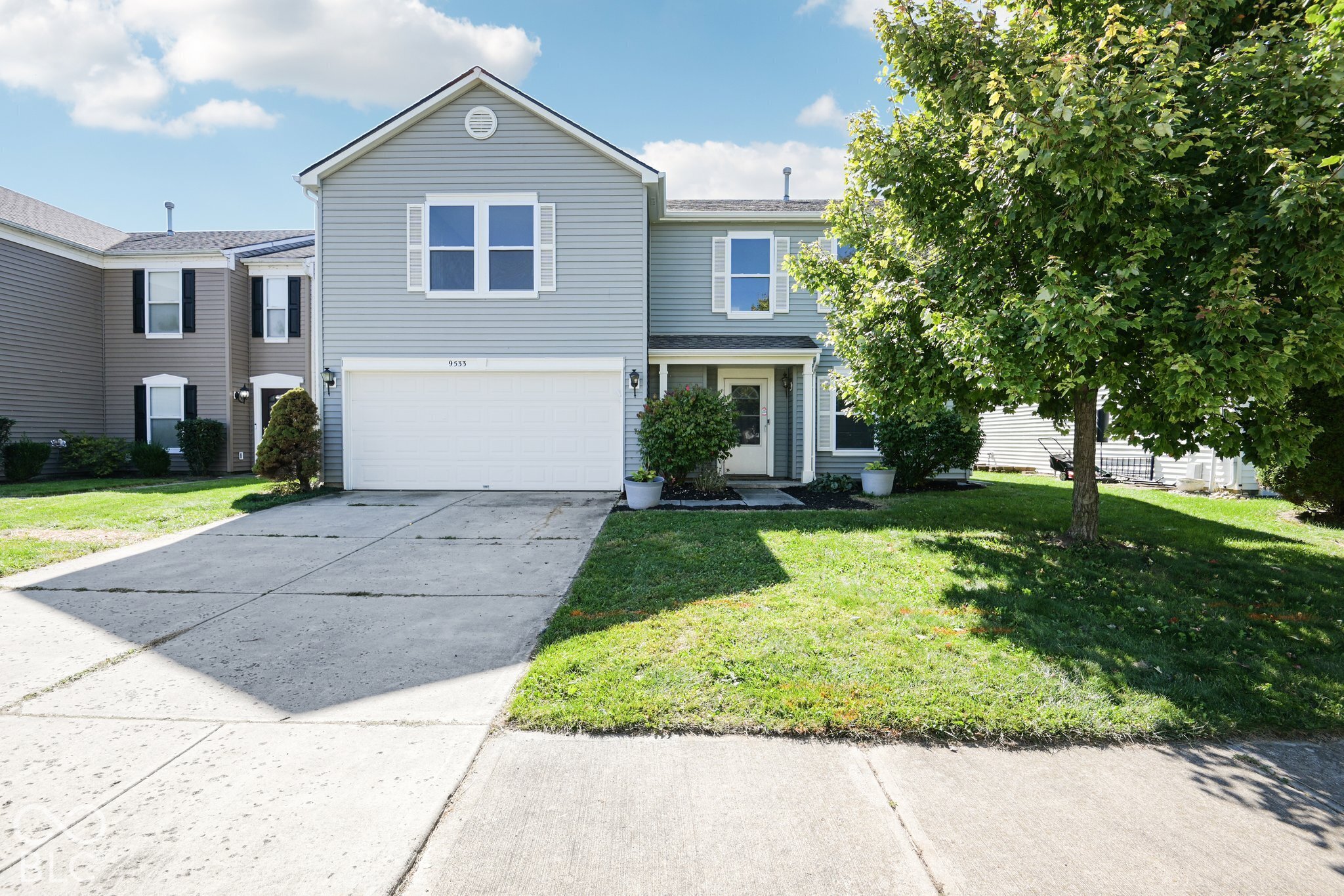 Gray two-story house with a double garage, broad driveway, and large leafy tree in the front yard.