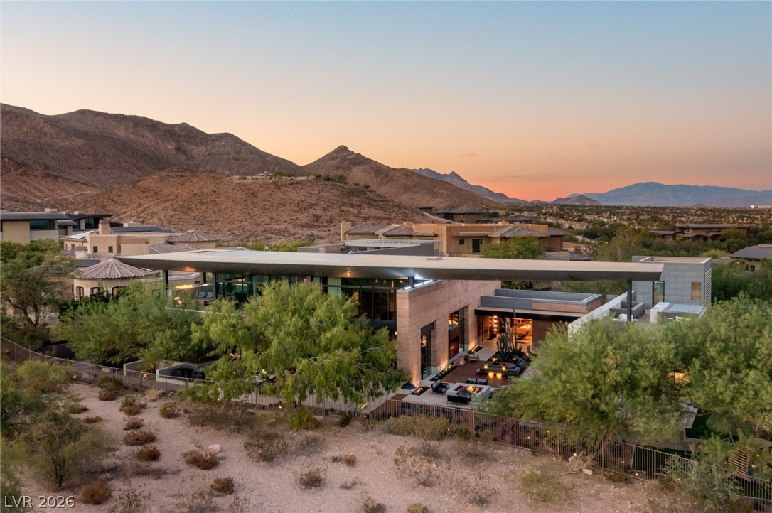 Modern desert home with an illuminated patio, surrounded by desert landscaping and mountains at dusk.