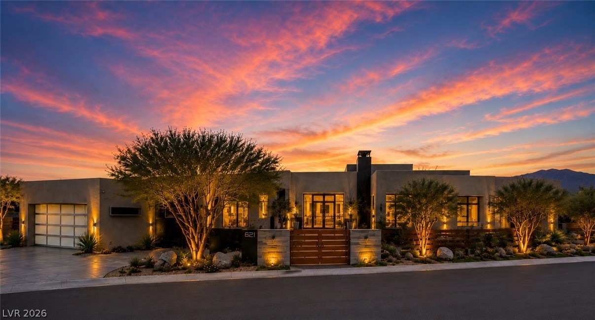 Modern desert home with a gated entrance and illuminated landscaping beneath a vivid sunset sky.