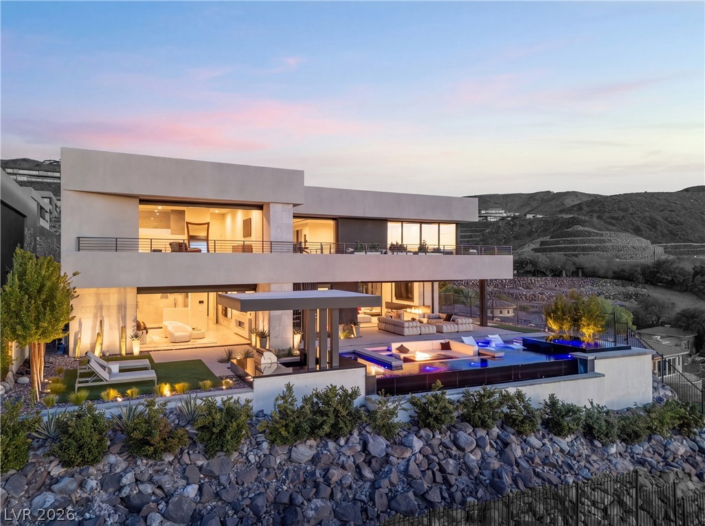 Modern two-story home with a tiered pool, illuminated balconies, and desert hills at dusk.