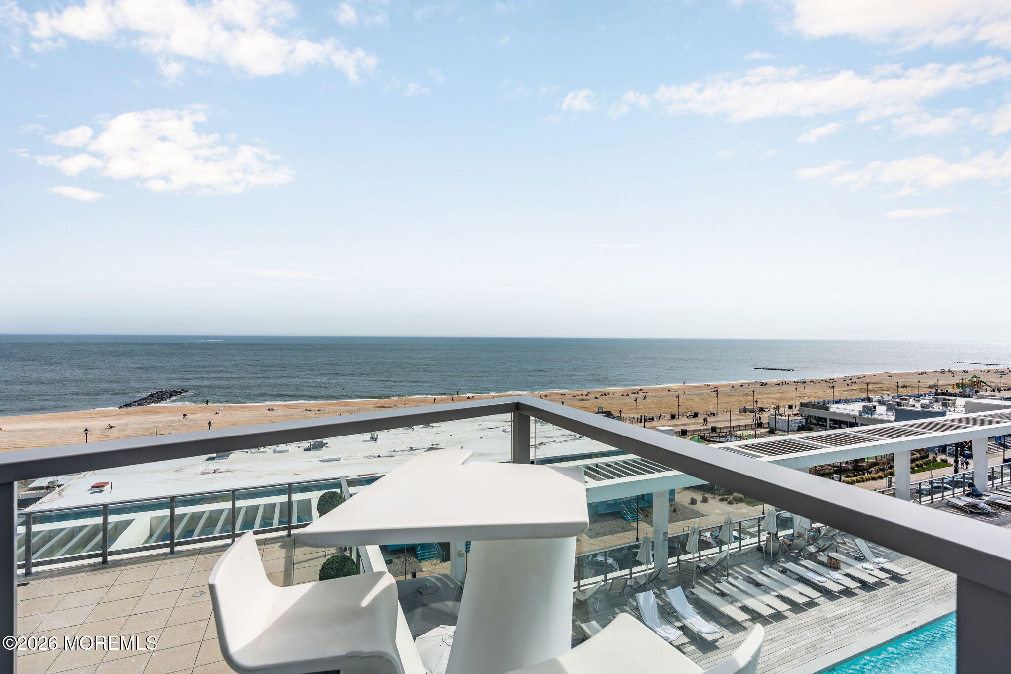 Oceanfront balcony overlooking the beach, with white patio furniture and a rooftop pool below.