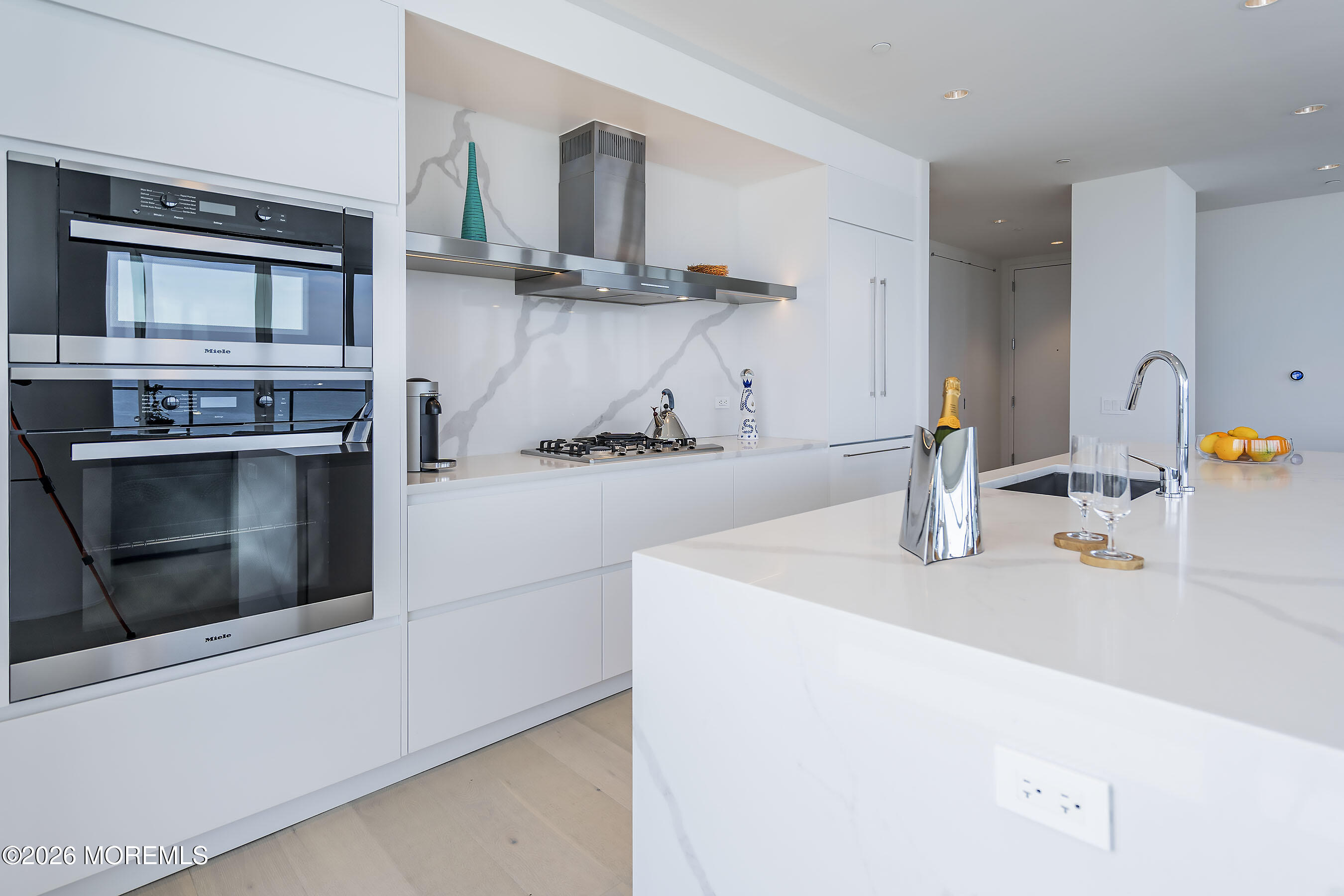 Modern white kitchen with a large quartz island, marble-veined backsplash, and stacked Miele ovens.