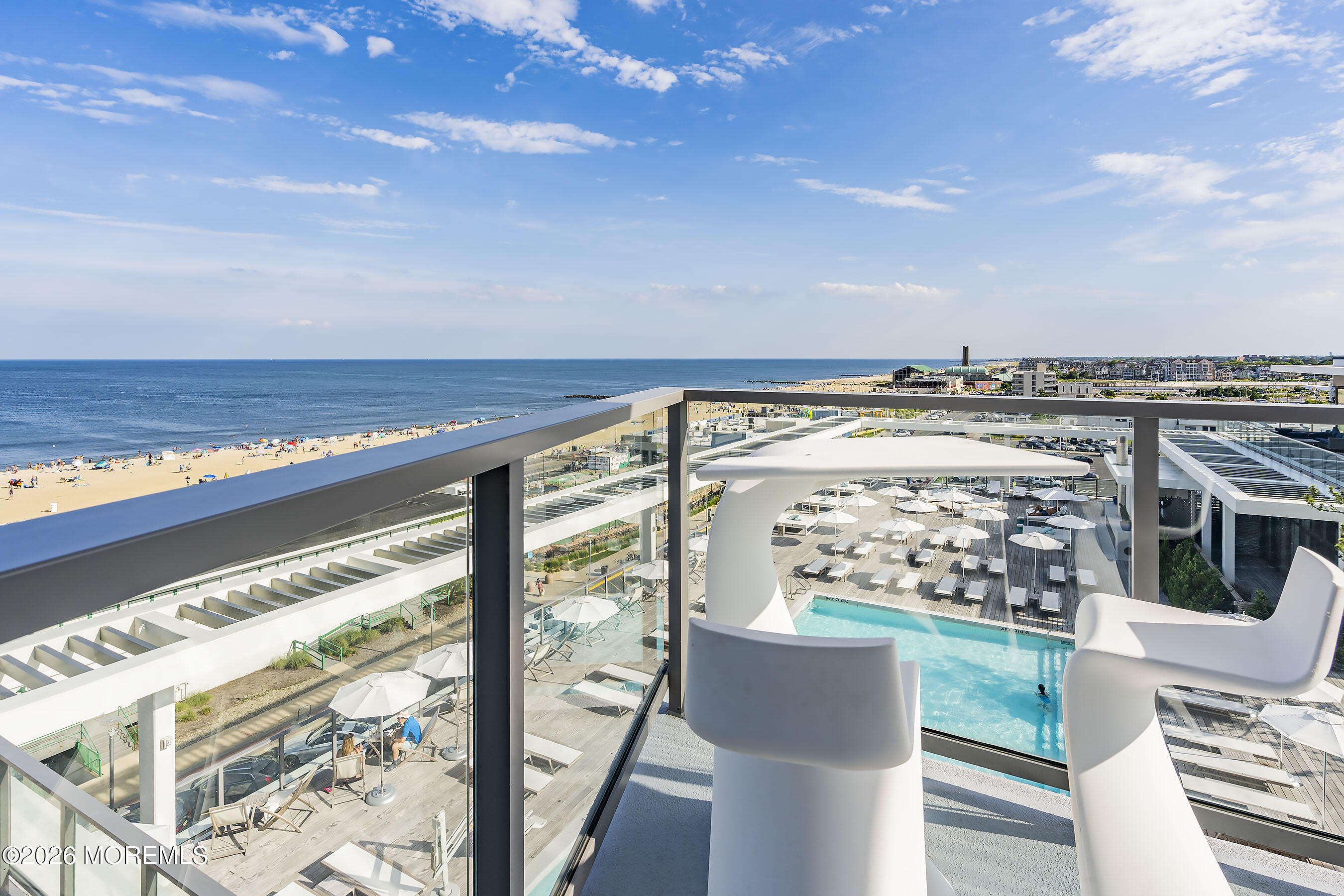 High-rise balcony overlooks the ocean and beach, with a pool terrace and white chairs below.