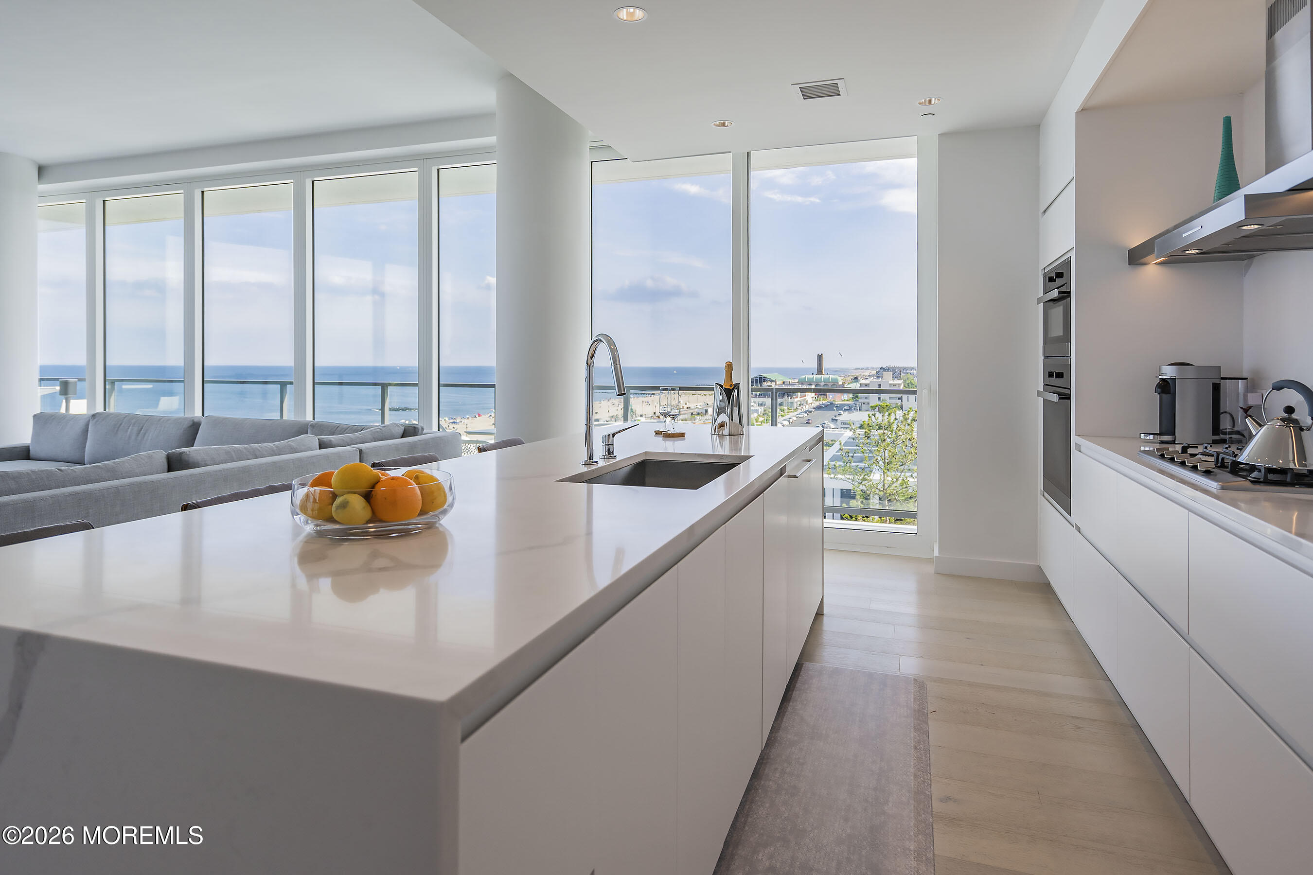 Modern white kitchen with a long island and floor-to-ceiling windows overlooking the ocean and beach.
