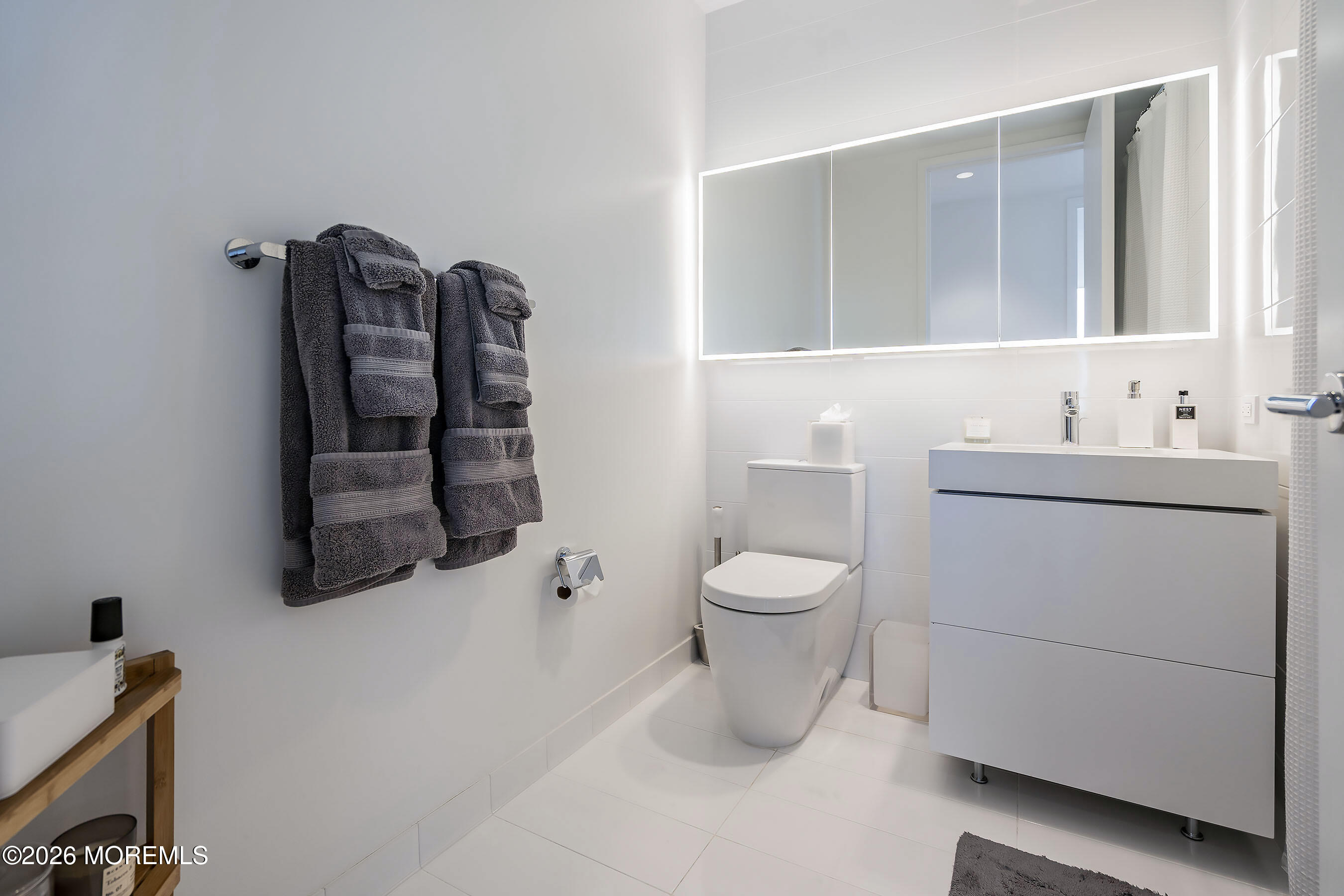 Modern bathroom with an illuminated mirror, gray vanity, white tile, and charcoal towels.
