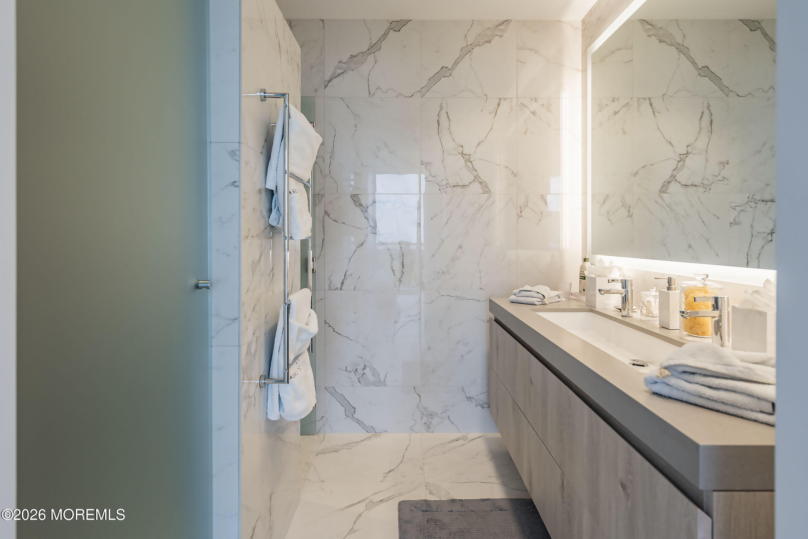 Marble-tiled bathroom with a glass shower and a long vanity topped with toiletries and folded towels.