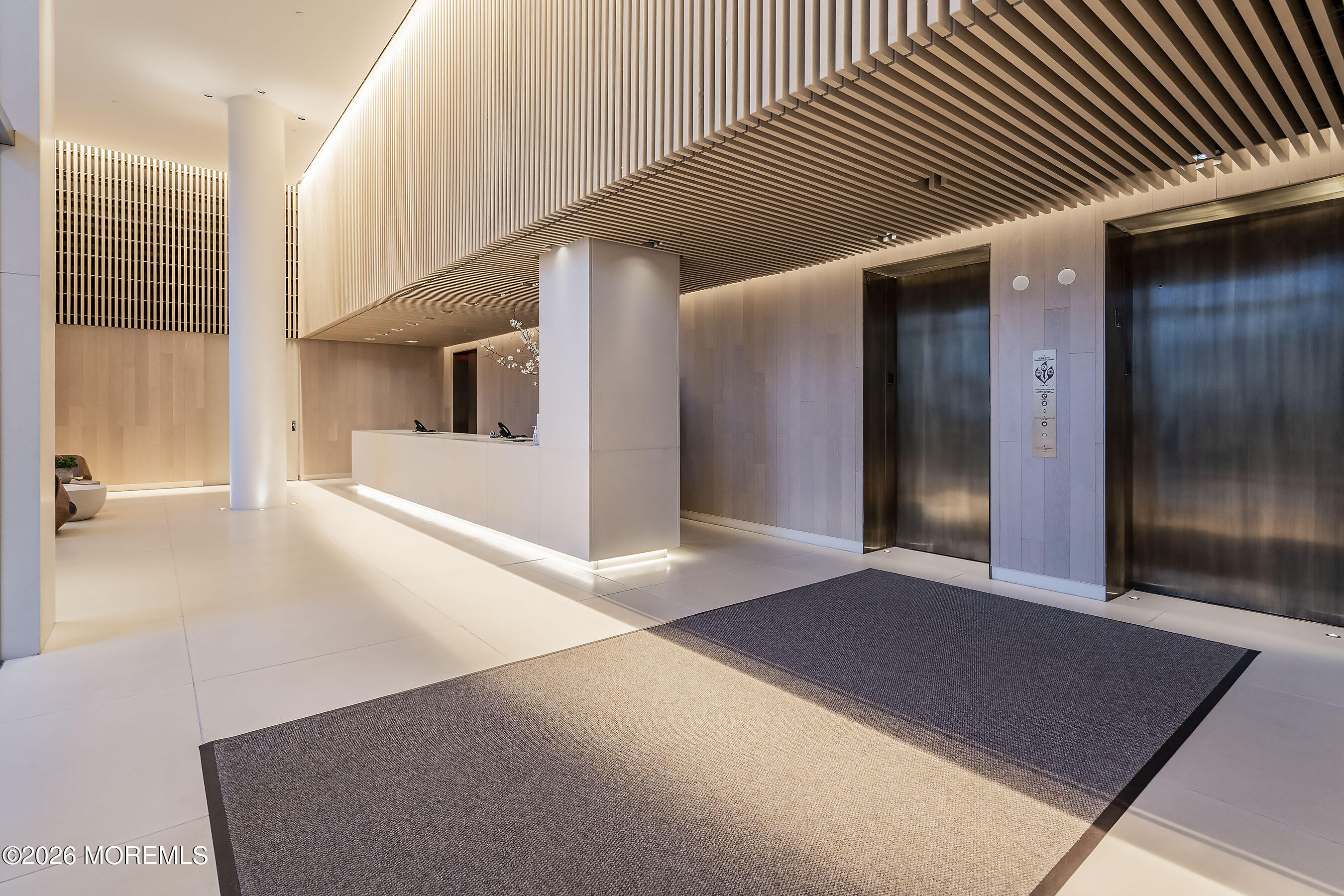 Modern condominium lobby with a reception desk, two elevators, and a dramatic slatted ceiling.