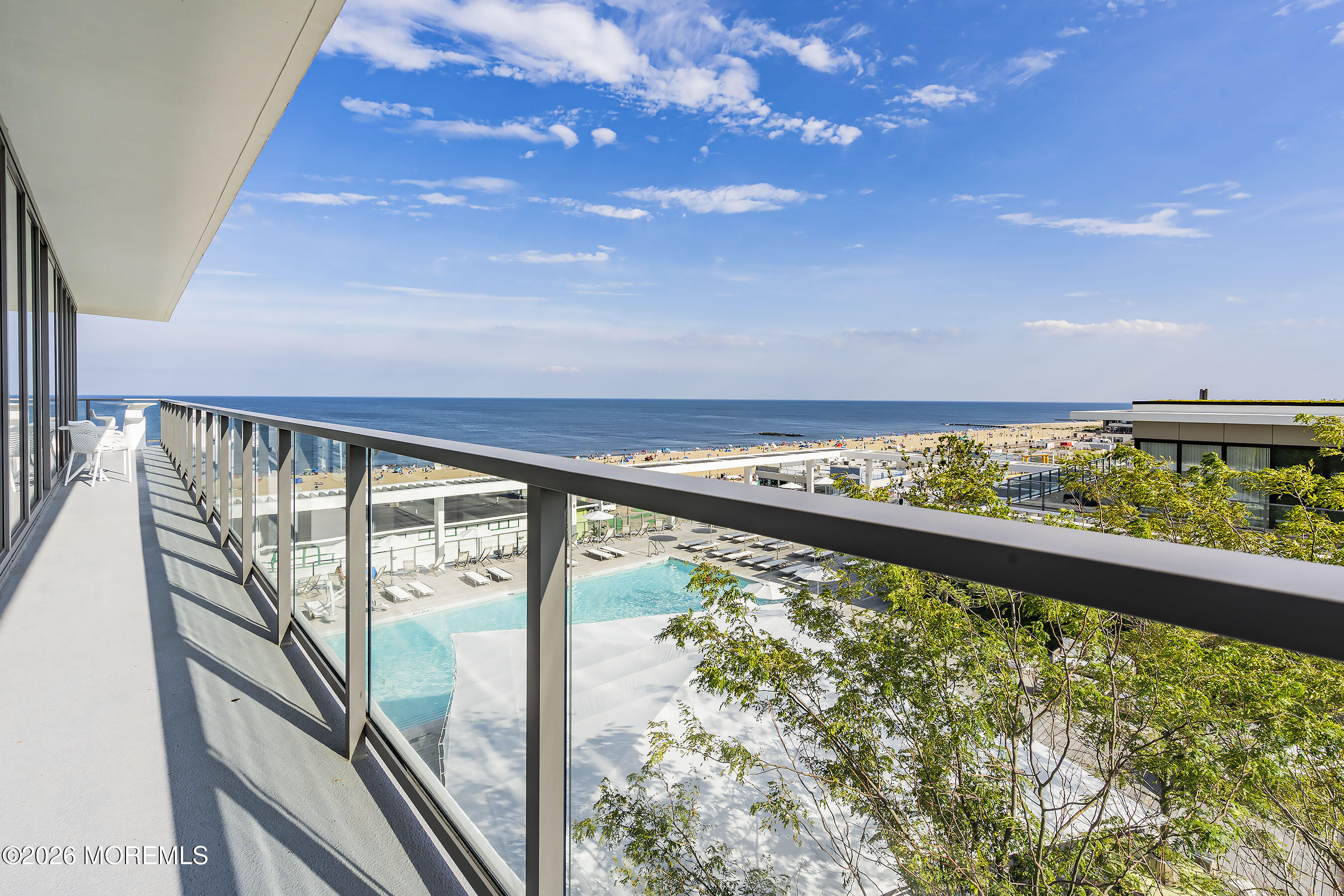 High-rise balcony overlooking a pool, sandy beach, and ocean, with glass railings and white patio chairs.
