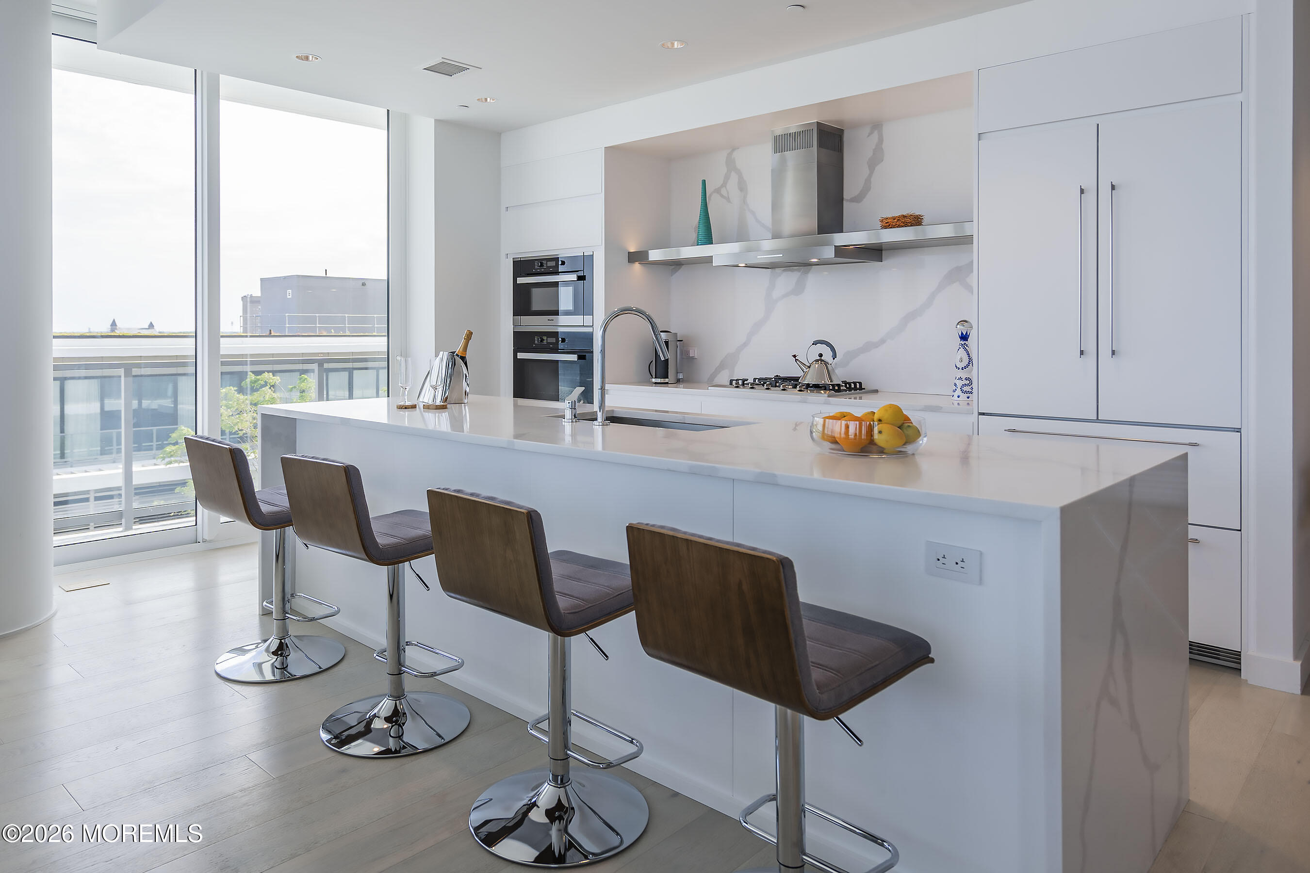 Modern white kitchen with a marble island, four bar stools, and floor-to-ceiling windows.