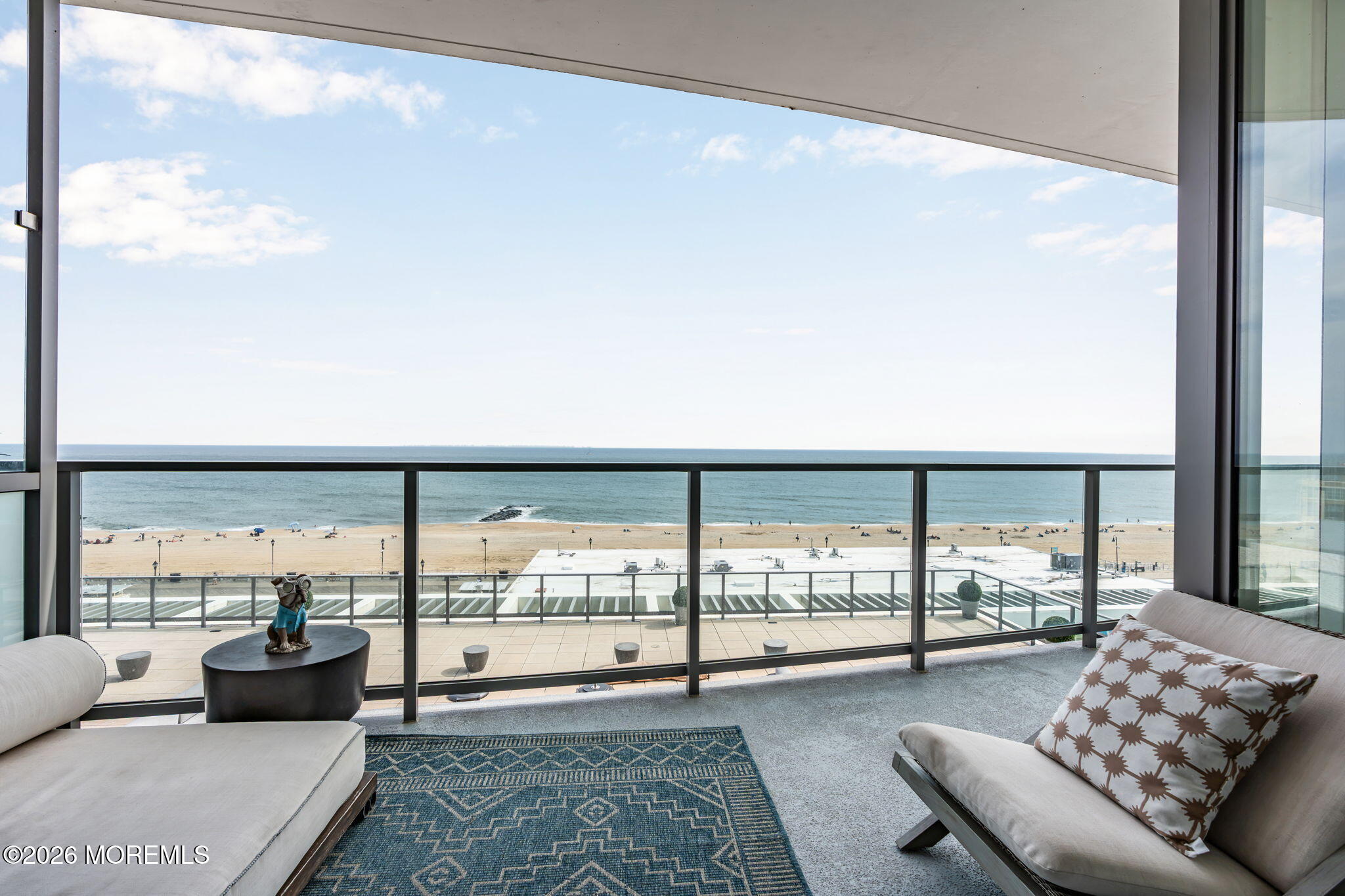Furnished living area and balcony overlooking the beach and ocean, framed by glass walls and lounge chairs.