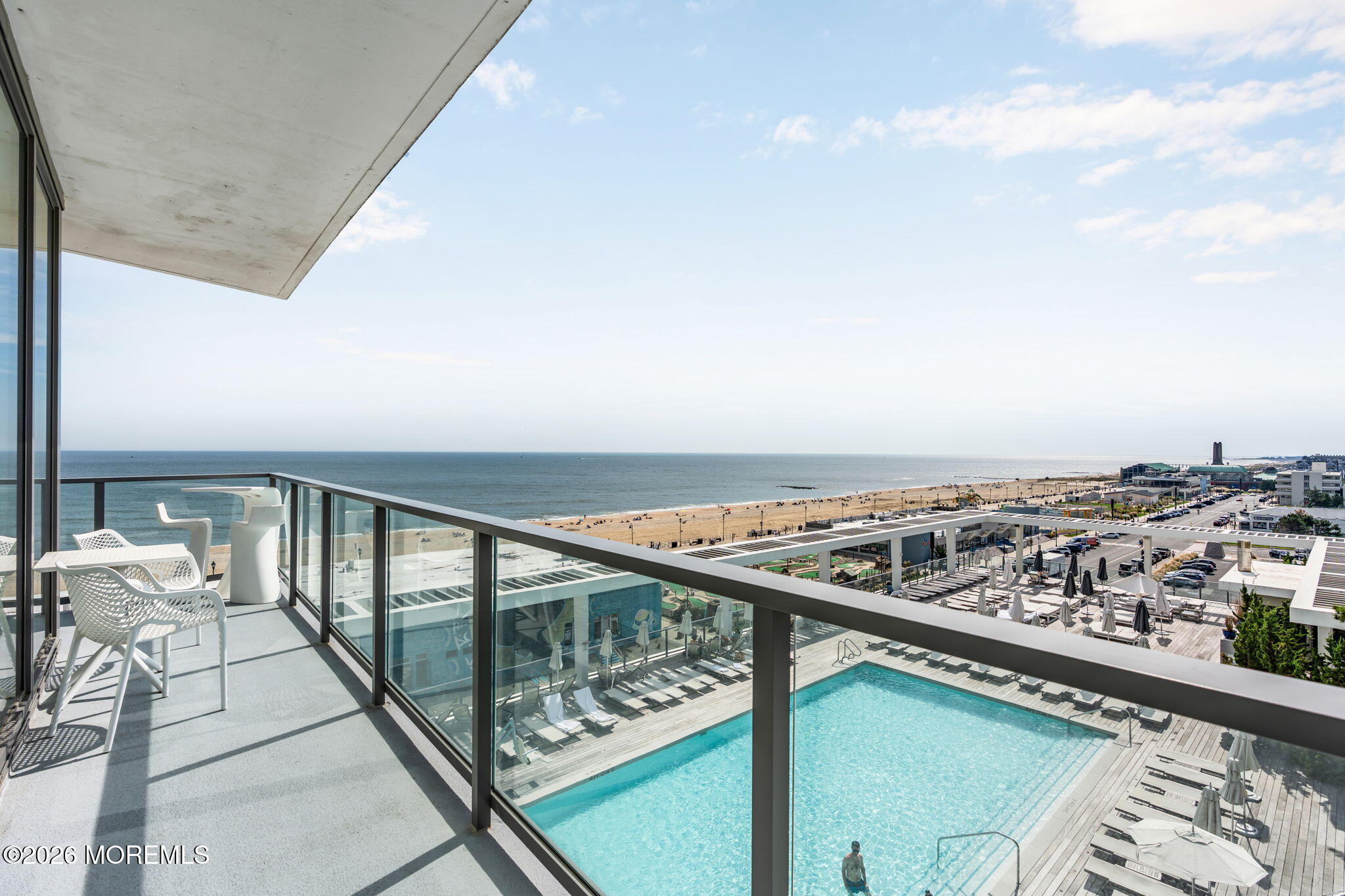 Oceanfront balcony with white chairs overlooks a swimming pool, sandy beach, and ocean.