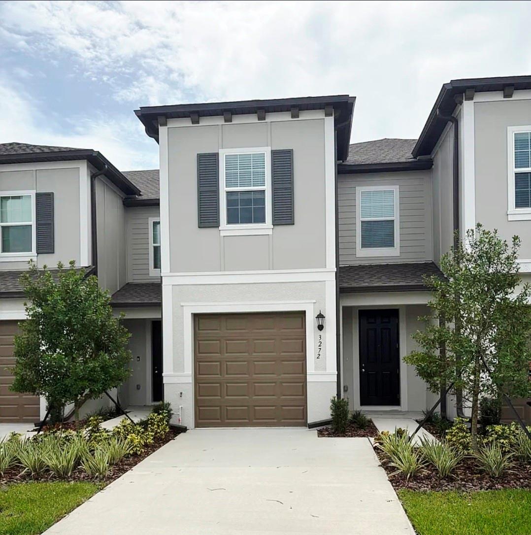 Gray two-story townhouse with a brown garage door, black entry door, and landscaped front beds.