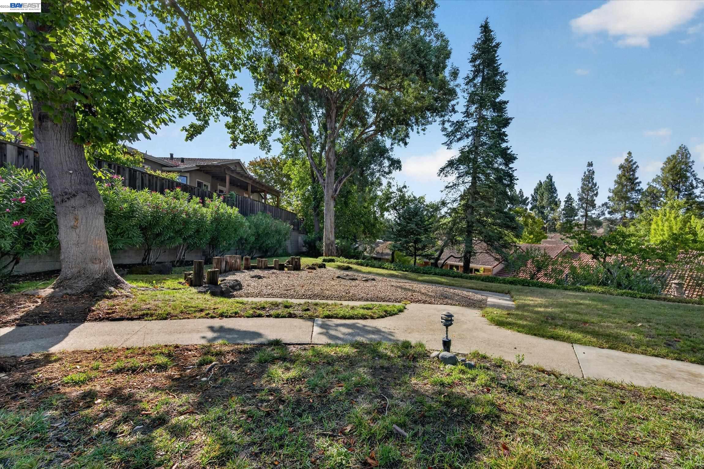 Landscaped backyard with mature trees, a gravel seating area, cut-log stools, and a curved concrete path.