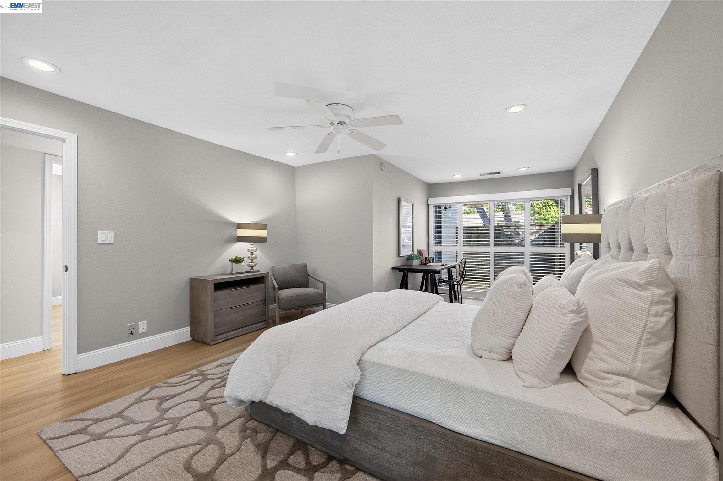 Furnished bedroom with a large upholstered bed, ceiling fan, and wide windows with blinds.
