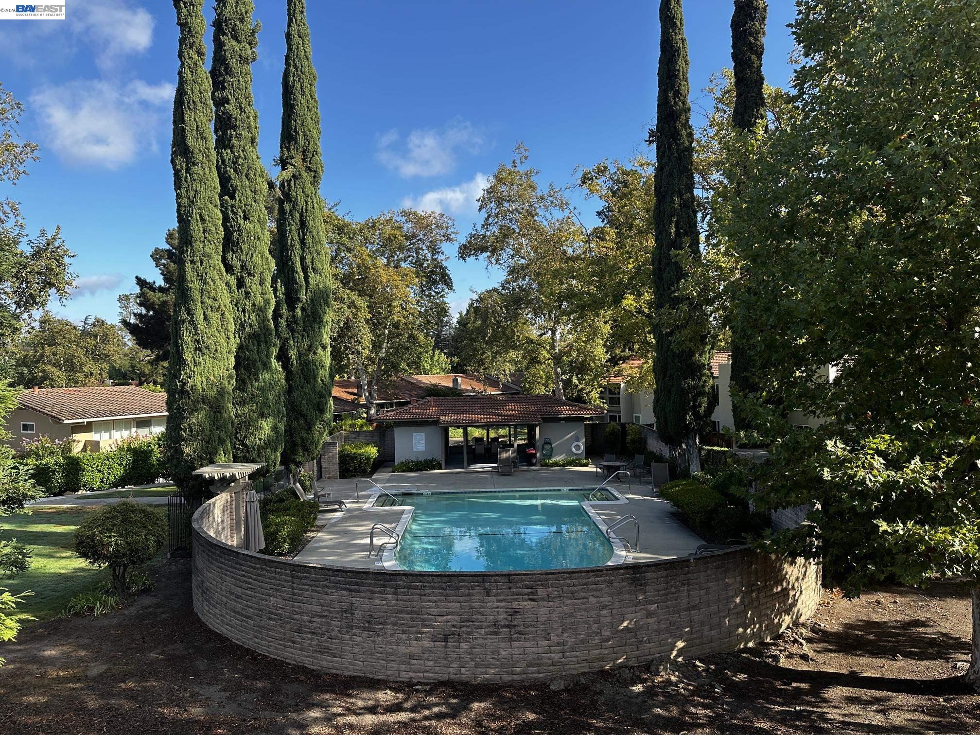 Outdoor swimming pool behind a curved brick wall, surrounded by cypress trees and patio seating.