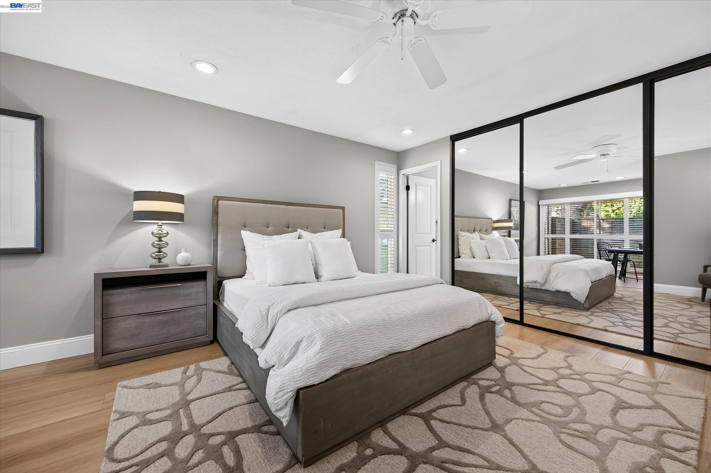 Staged bedroom with a white bed, mirrored closet doors, and a large patterned rug.