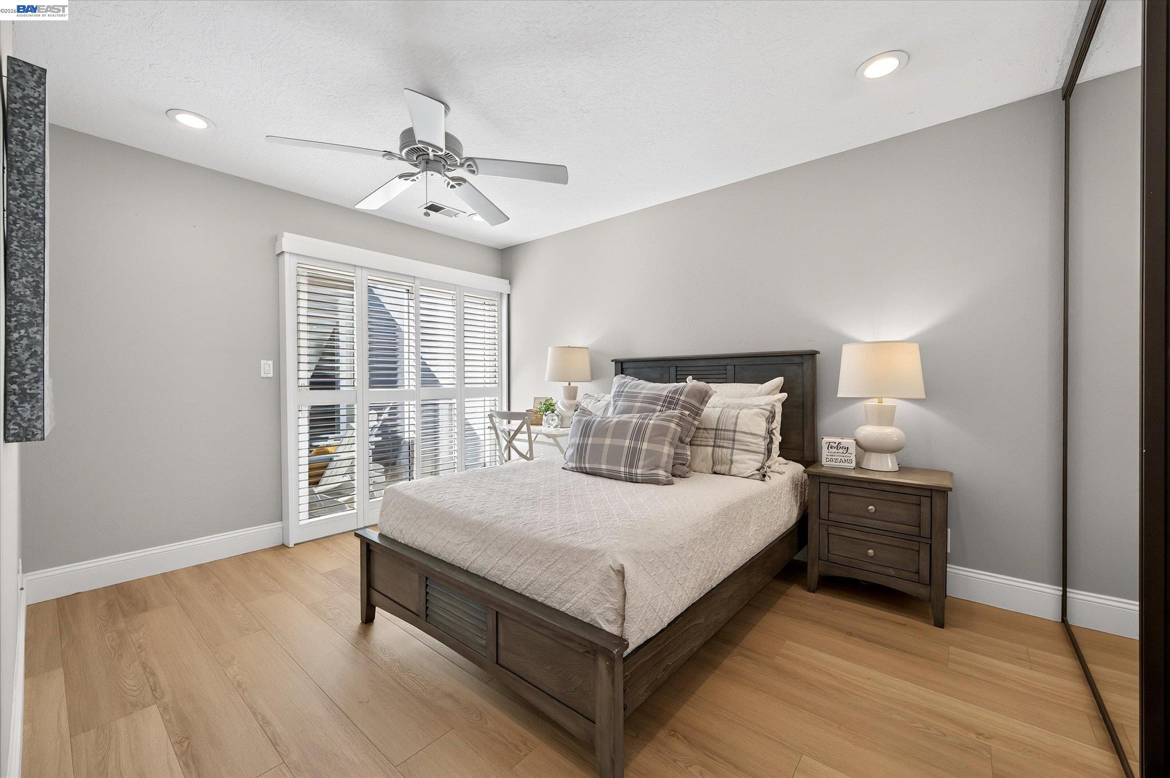 Bedroom with a dark wood bed, plaid pillows, plantation shutters, and light wood flooring.