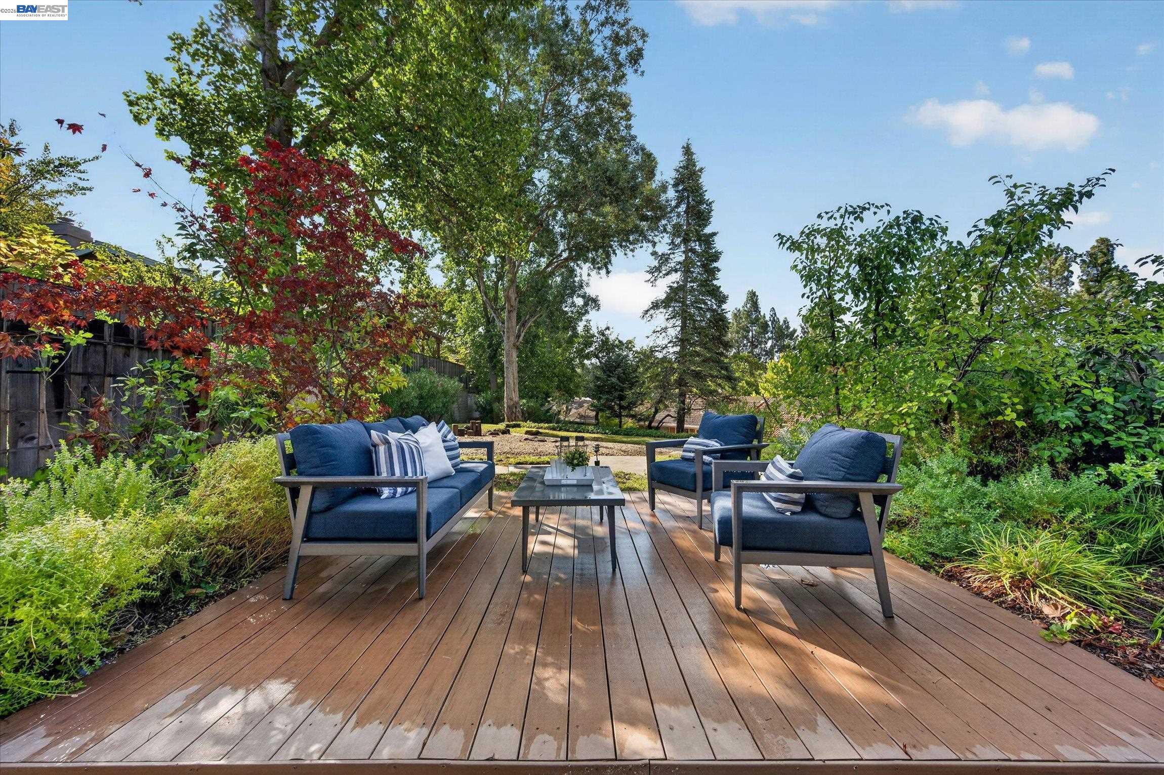Wooden backyard deck with navy seating and a coffee table, surrounded by mature trees and greenery.