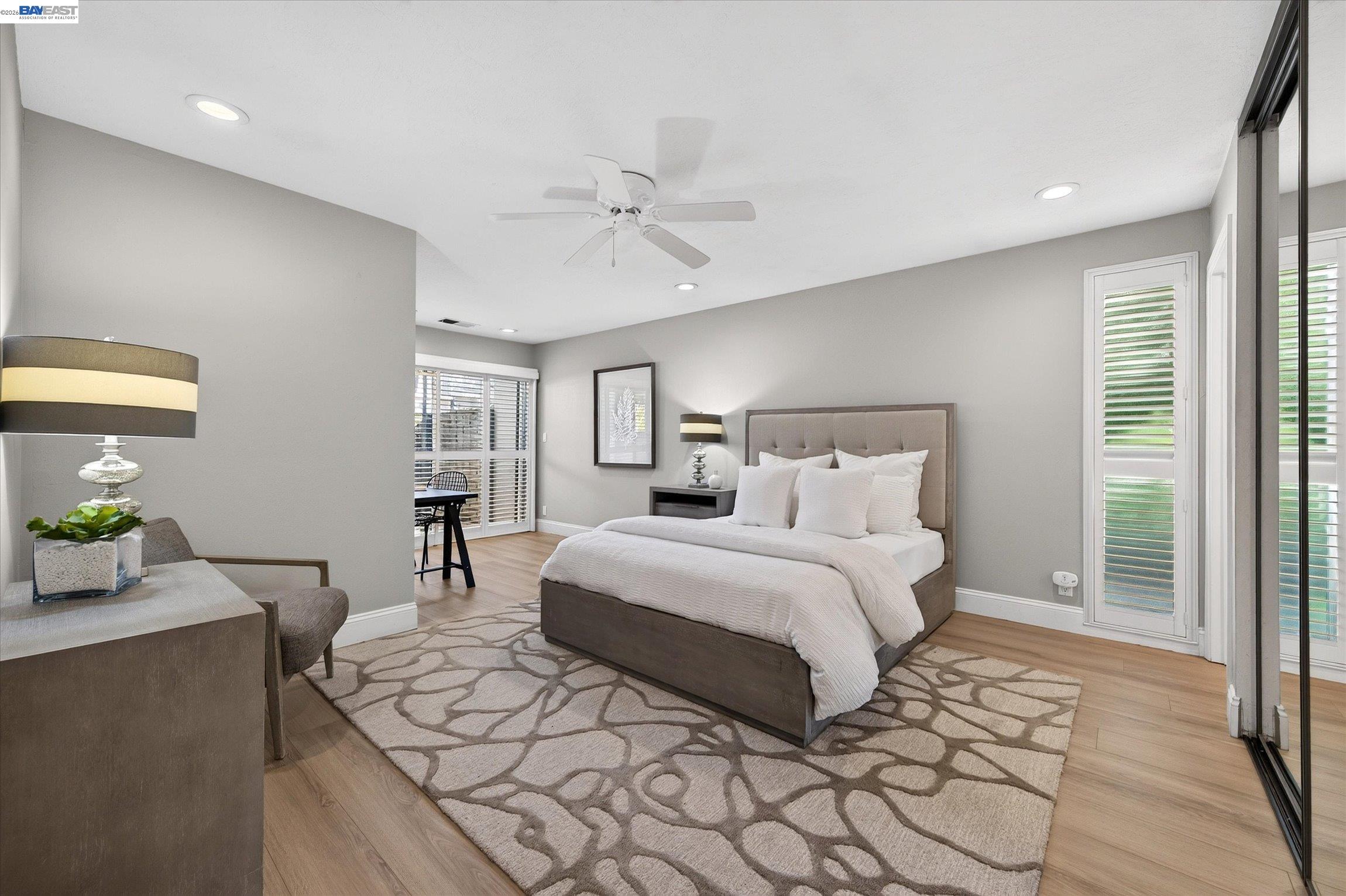 Staged bedroom with a white bed, patterned rug, ceiling fan, and plantation shutters.