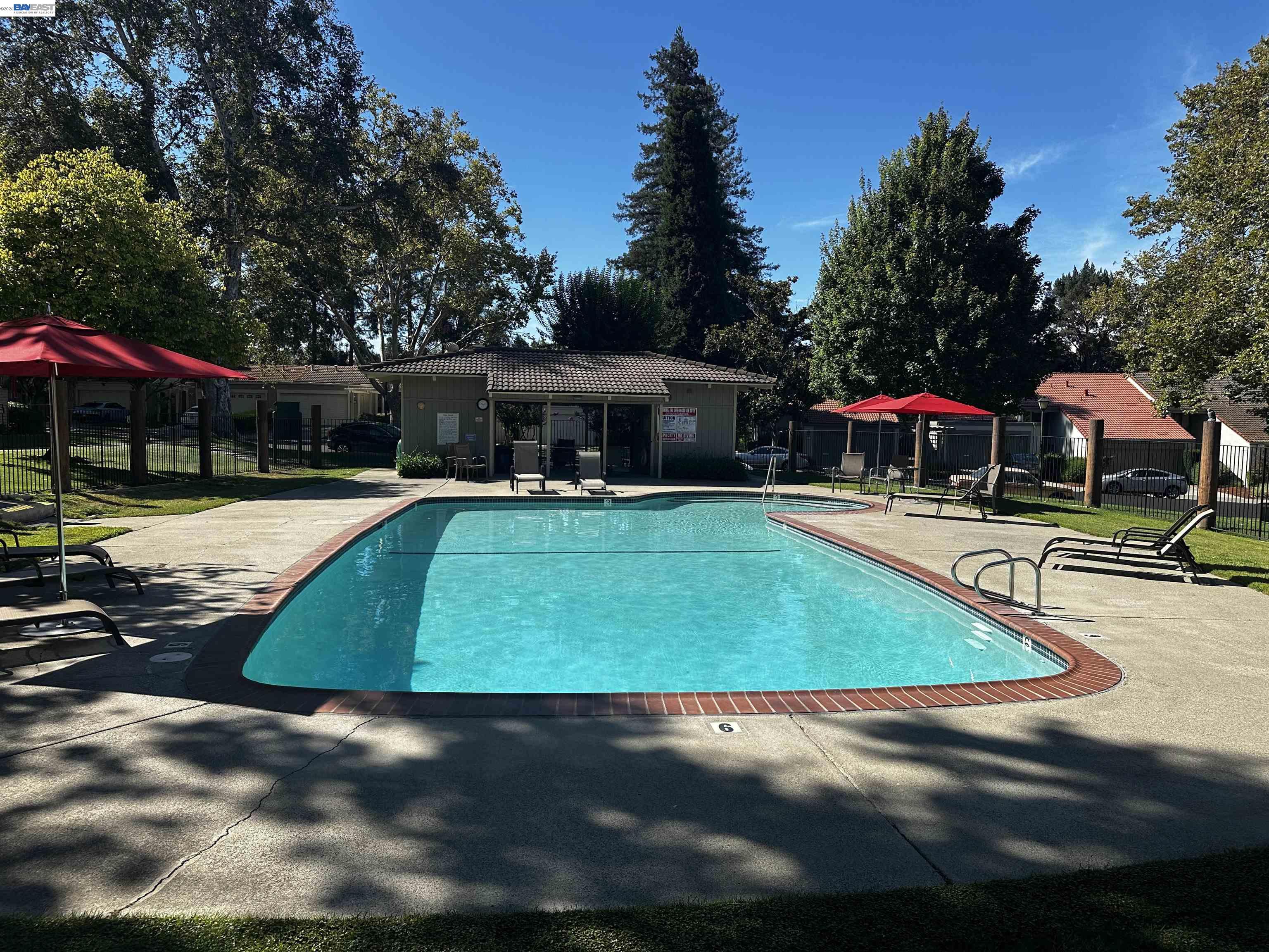 Community pool with brick edging, lounge chairs, red umbrellas, and mature trees.
