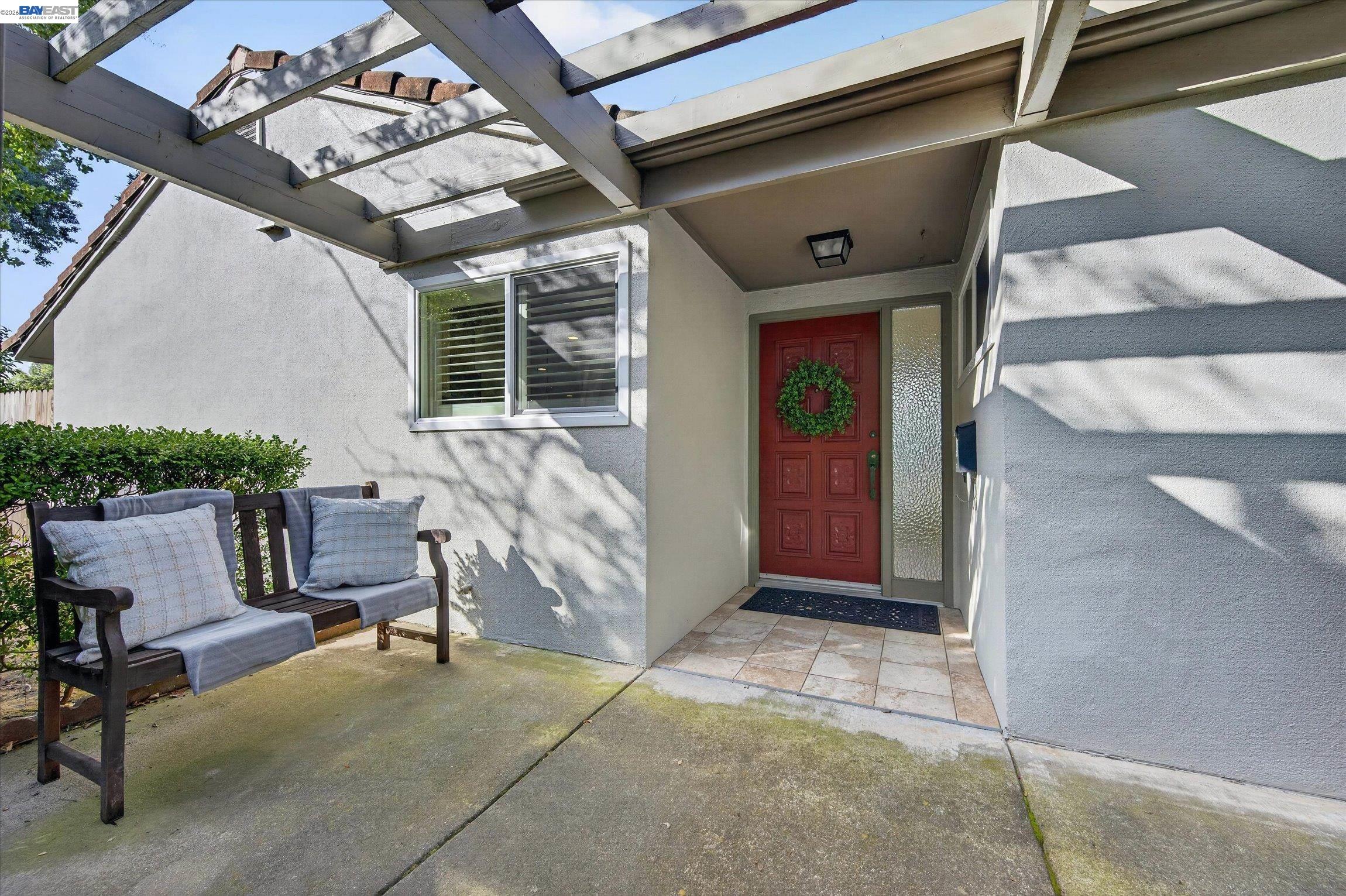 Covered home entry with a red wreath-adorned door, tiled landing, and cushioned bench beneath a pergola.