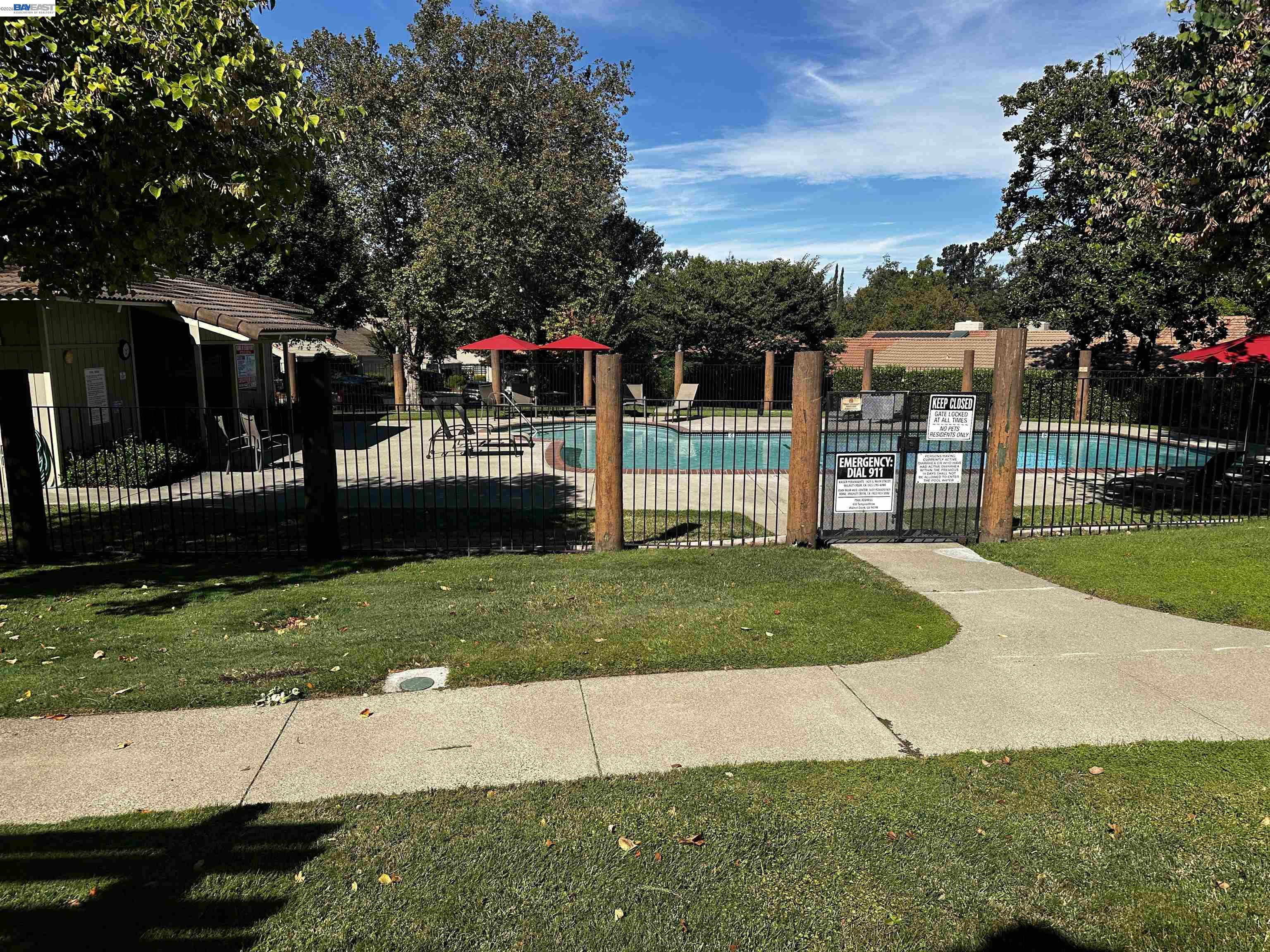 Fenced community swimming pool with red umbrellas, lounge chairs, and mature trees.