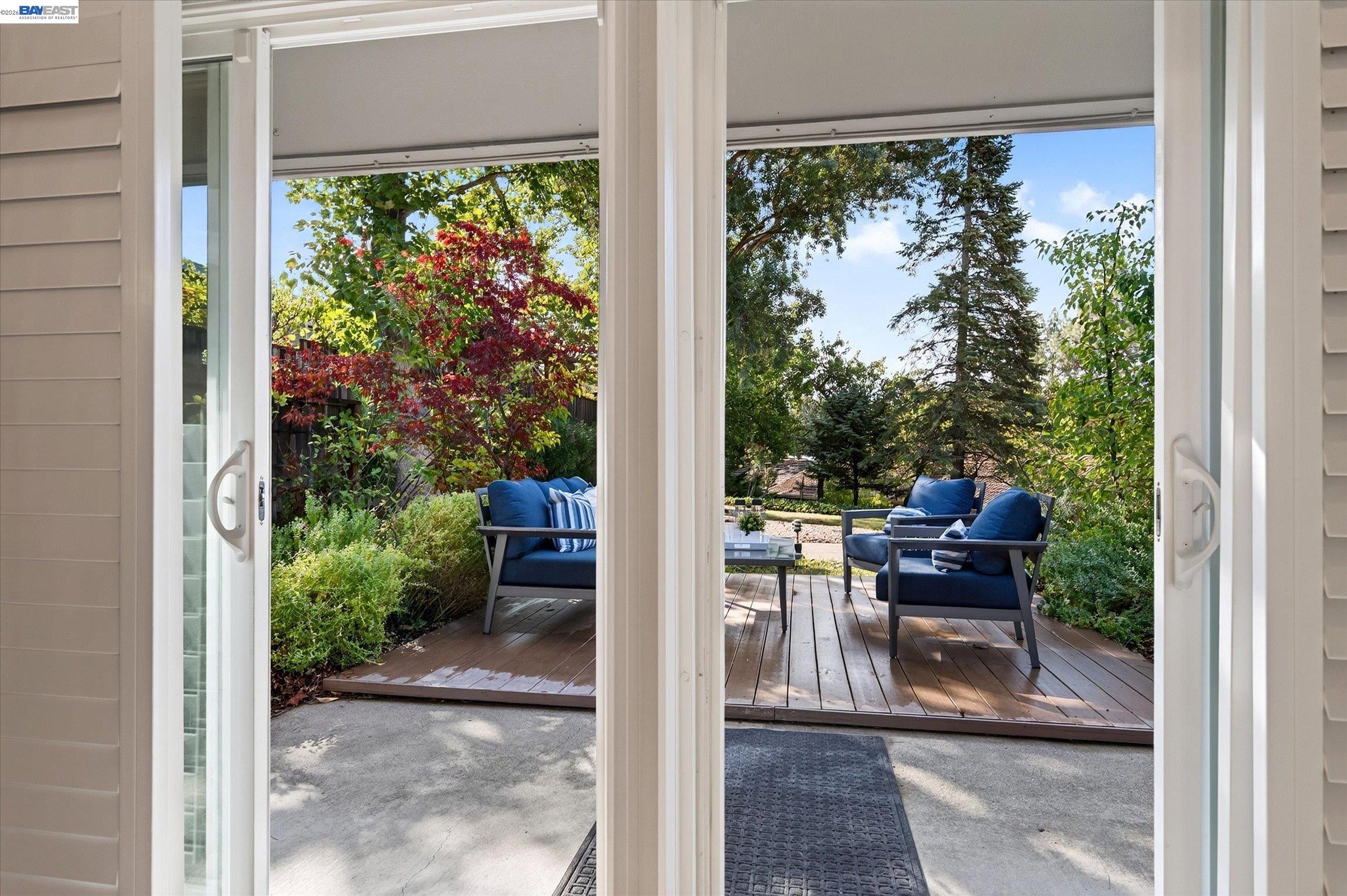 Wood deck patio with blue-cushioned seating, framed by open sliding doors and leafy garden greenery.
