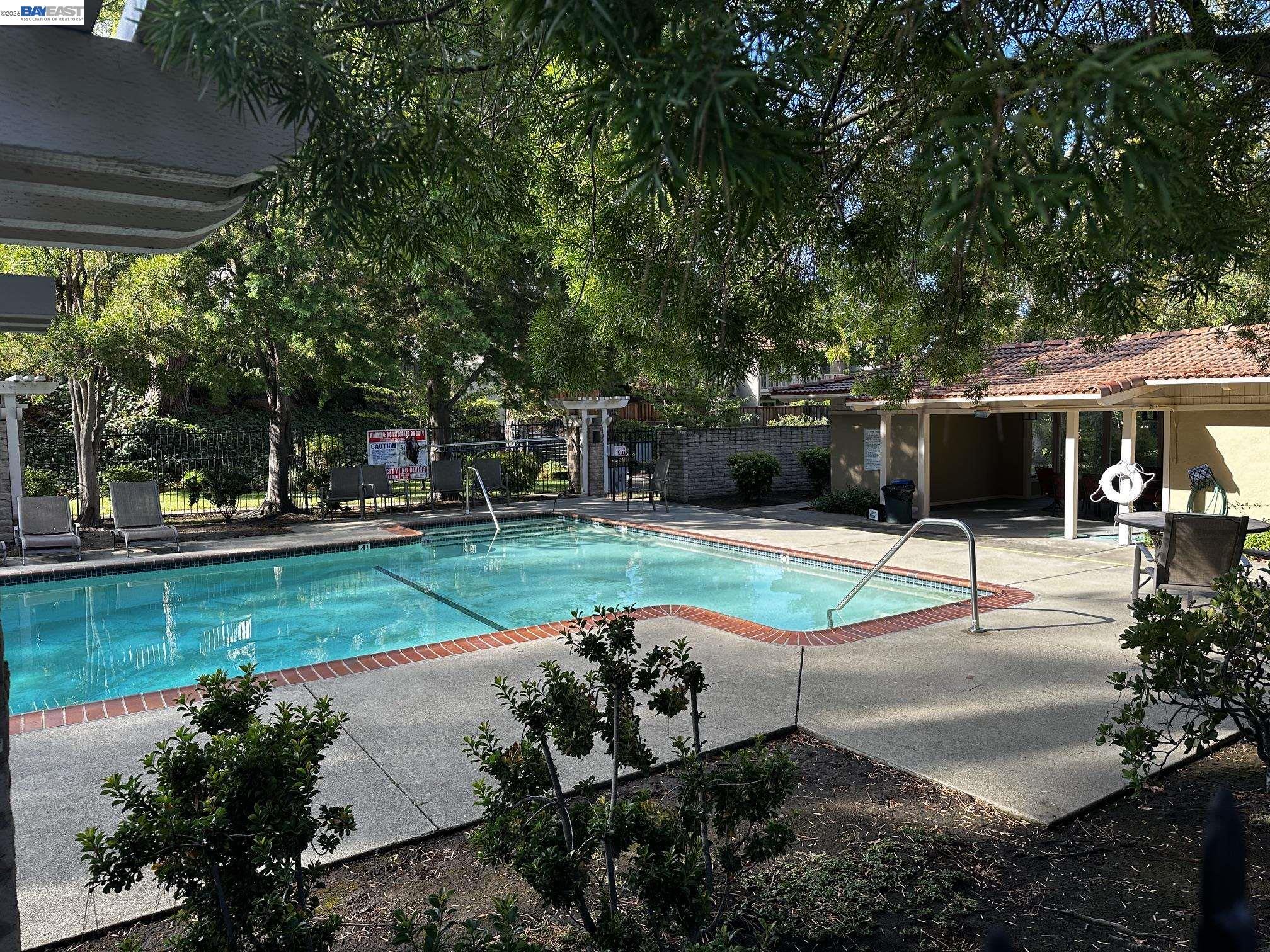 Outdoor swimming pool bordered by brick coping, shaded by mature trees beside a covered cabana.