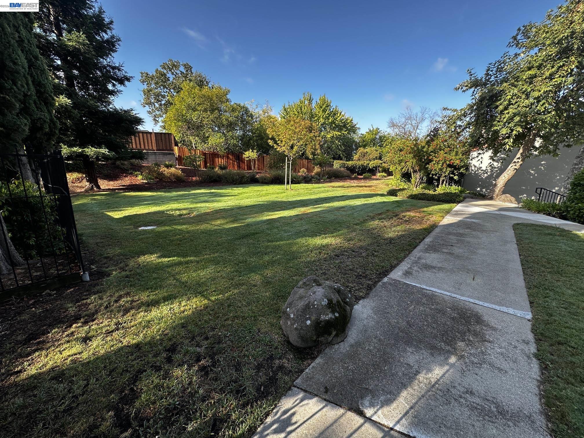 Backyard lawn bordered by mature trees and a wooden fence, with a concrete walkway and large rock.