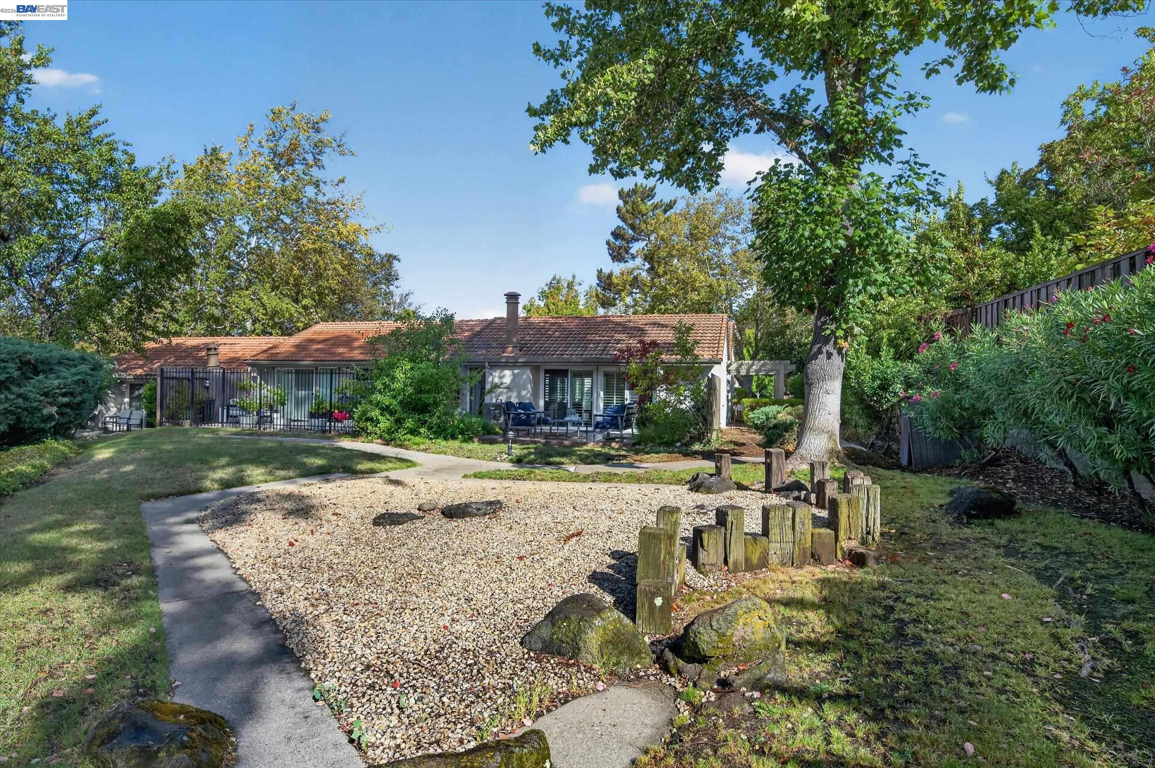 Single-story home viewed from the backyard, with a gravel area, mature trees, and patio seating.