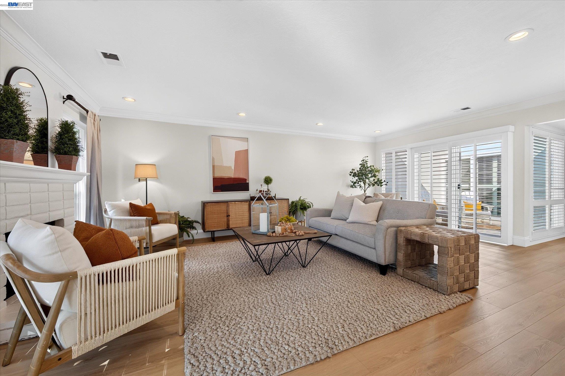 Staged living room with a white brick fireplace, gray sofa, woven chairs, and plantation shutters.