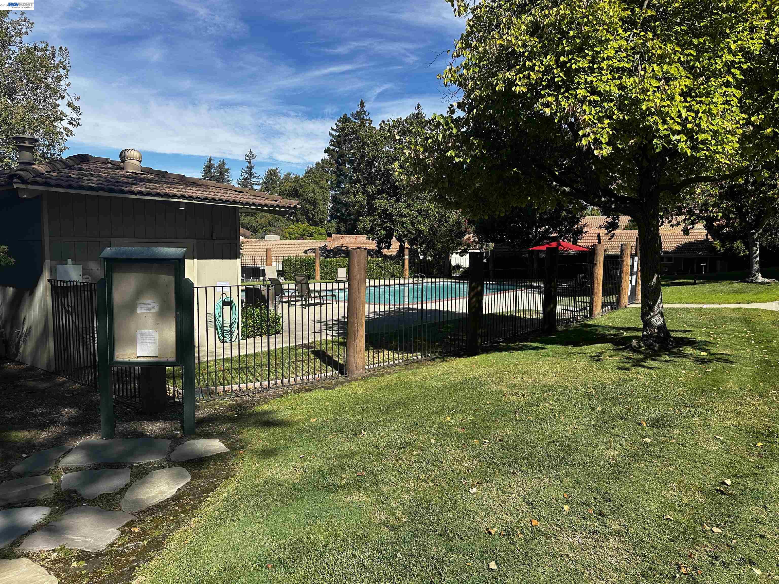 Community swimming pool behind a black fence, shaded by mature trees beside a grassy lawn.