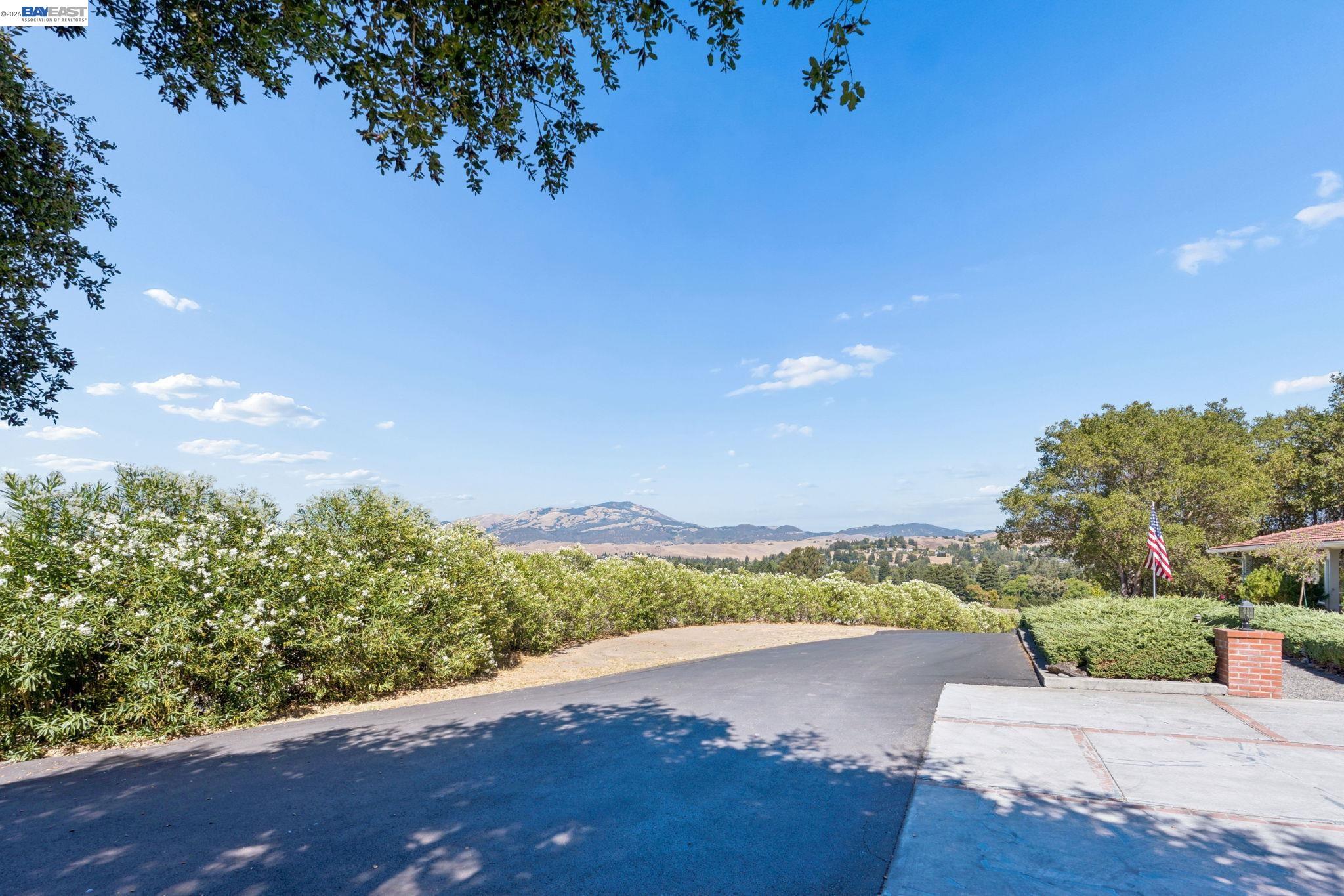Paved driveway and flowering hedges frame a distant mountain view; an American flag stands beside the home.