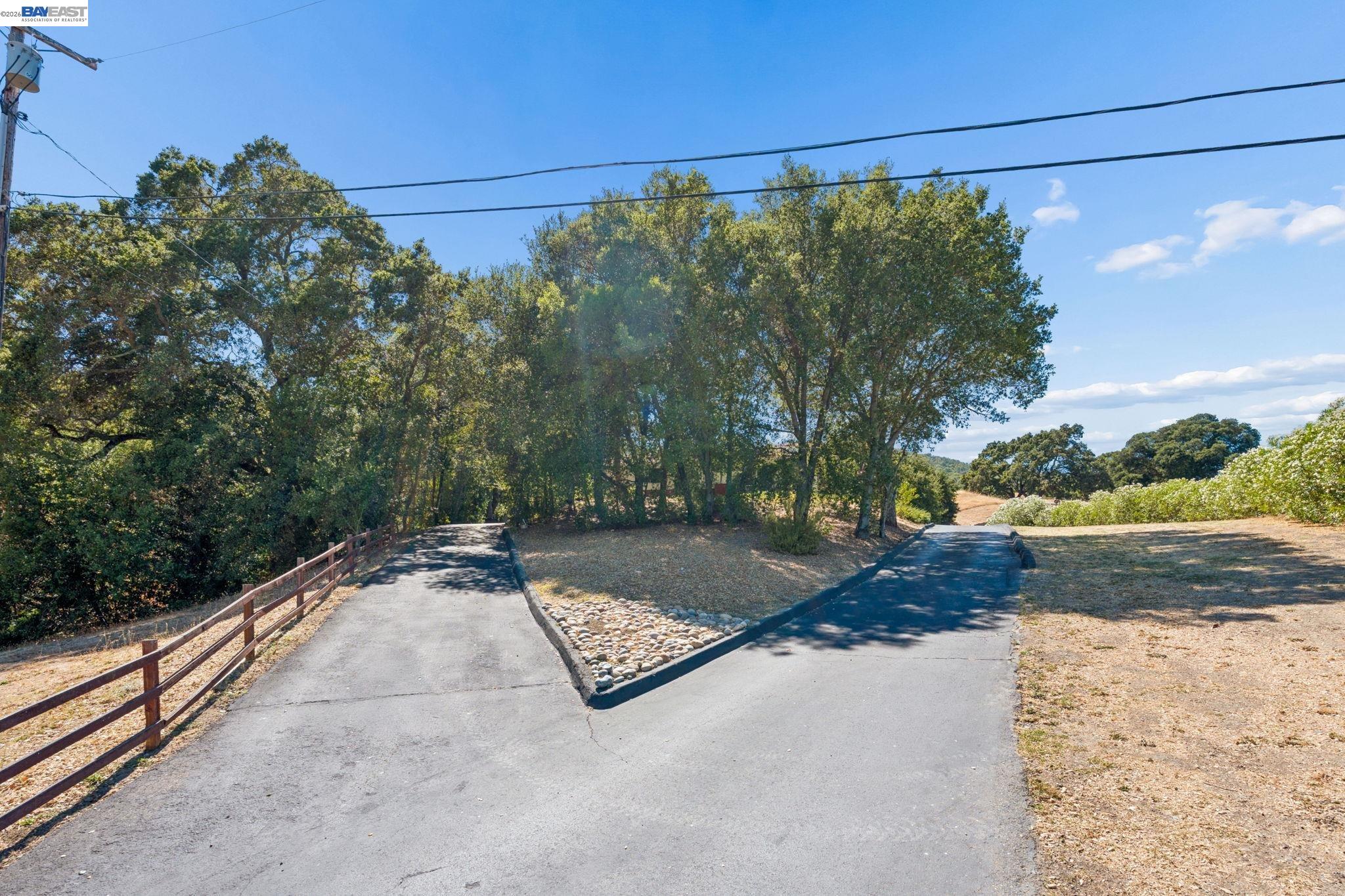 Paved driveway forks around a stone-edged island beneath mature trees, with a wooden rail fence along one side.