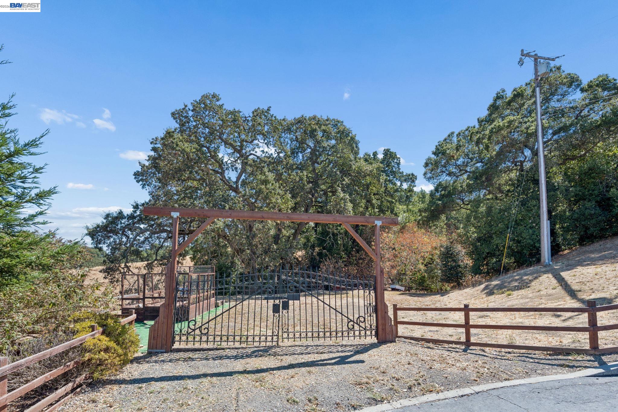 Property entrance with an ornate iron gate beneath a wooden arbor, surrounded by trees and fencing.