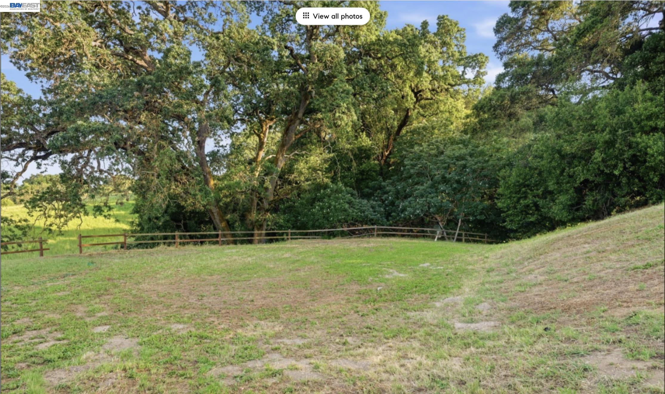Grassy sloping property bordered by mature trees and woodland, with a wooden rail fence.