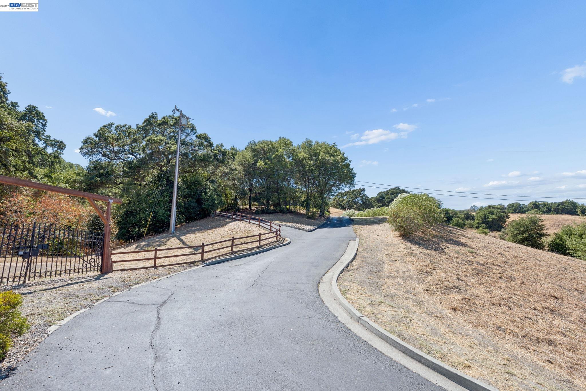 Paved access road curving past a metal gate and wooden fencing through dry, tree-lined land.