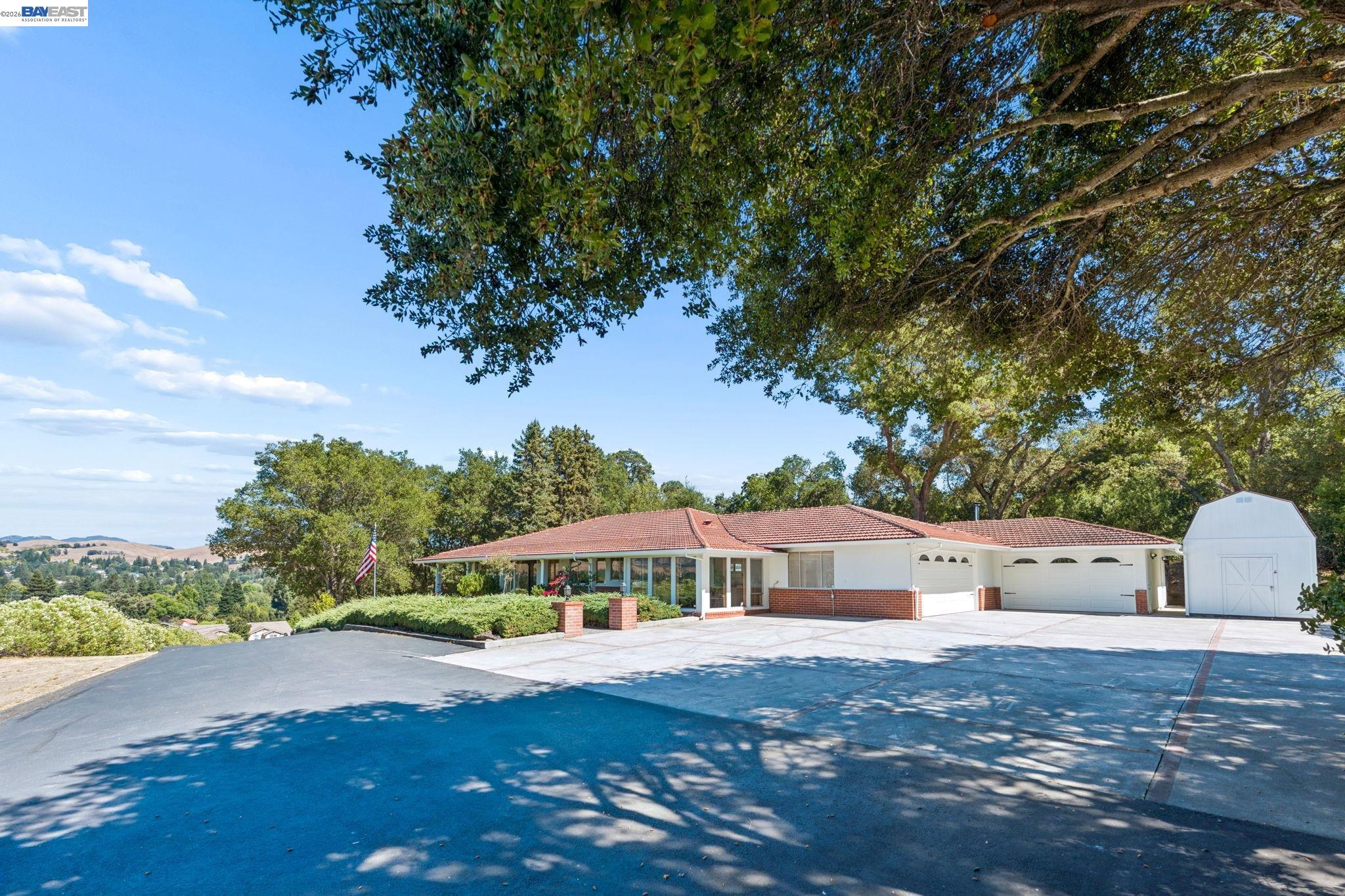 Single-story home with a red tile roof, broad driveway, attached garage, and detached white shed.