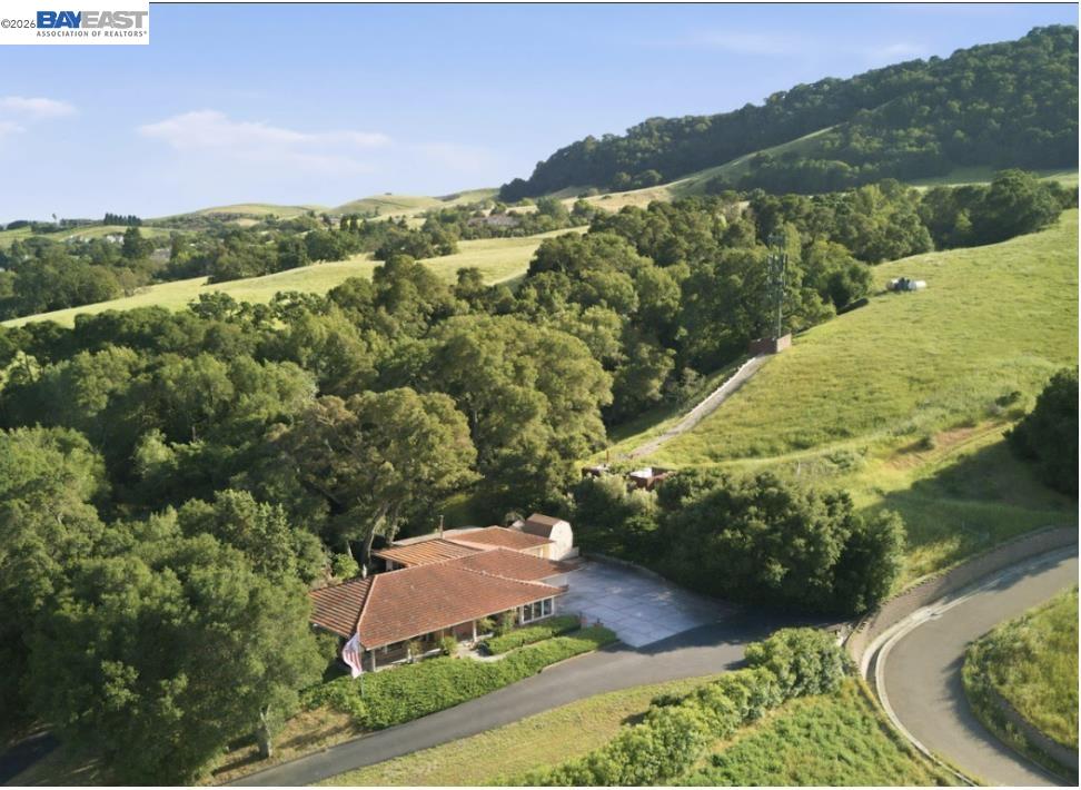 Aerial view of a red-tile-roof home amid wooded hills and rolling green fields.