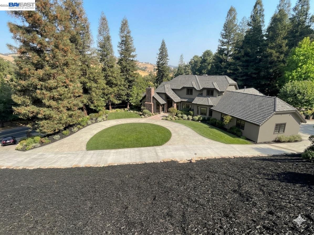 Large home with gray tiled roofs, circular driveway, landscaped lawn, and wooded hills behind it.