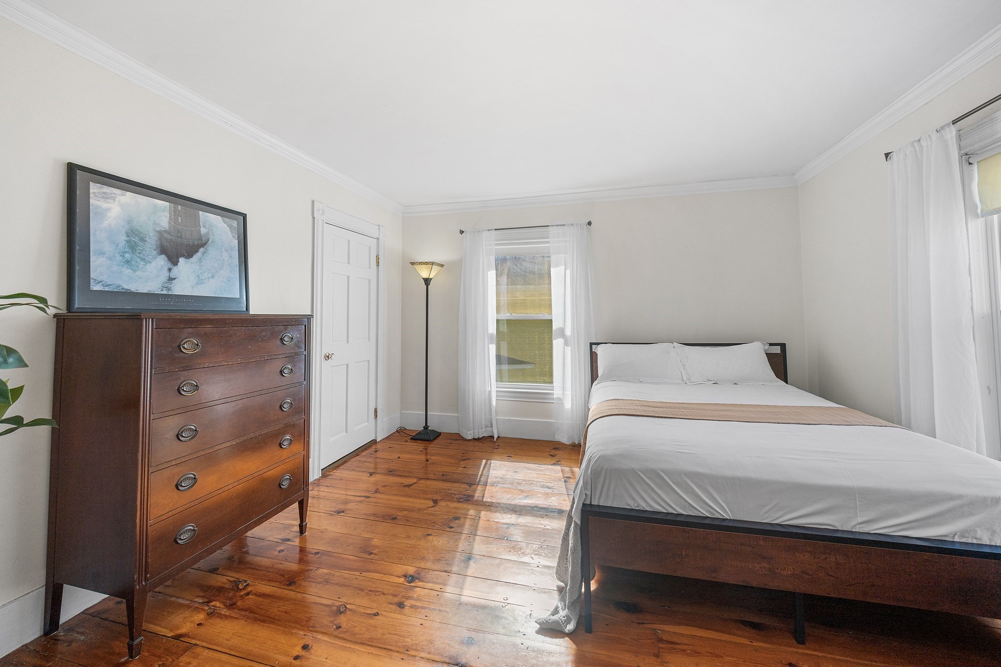 Bedroom with a dark wood bed, tall dresser, sheer curtains, and polished wide-plank floors.
