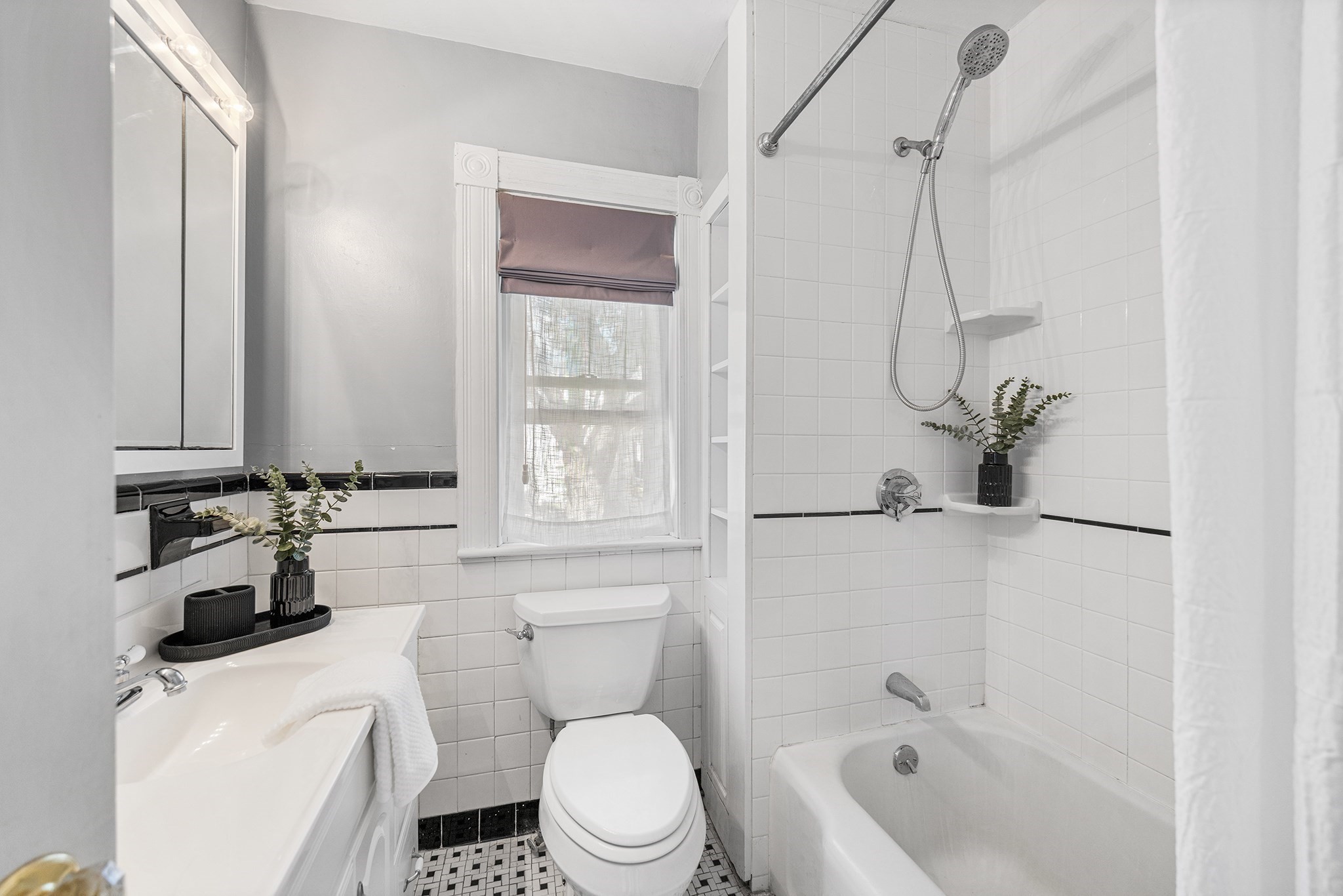 Bright tiled bathroom with a tub and shower, window, white vanity, and black tile accents.