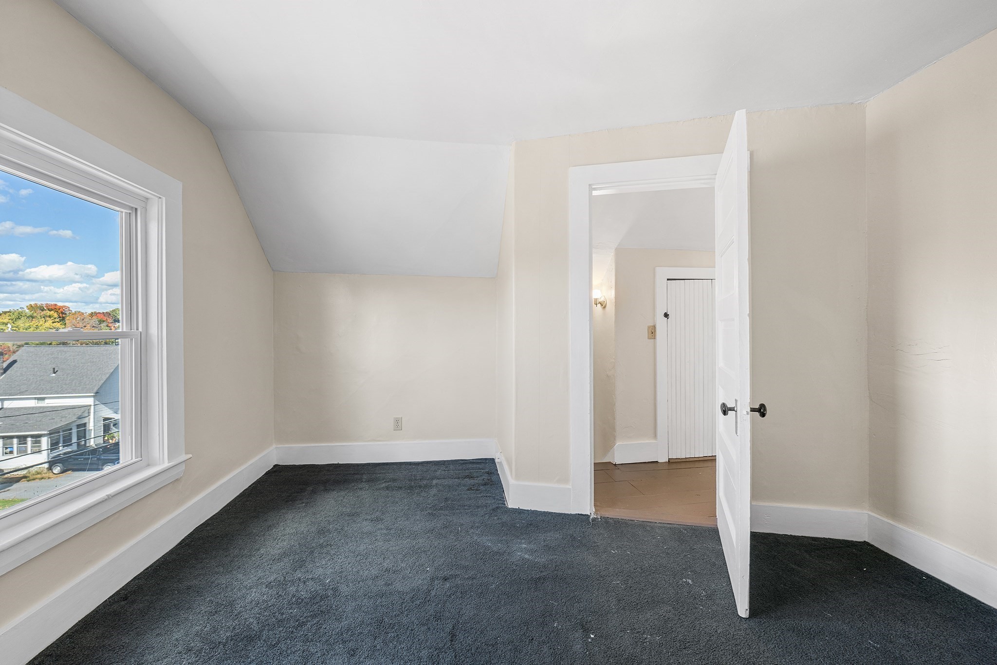 Empty bedroom with dark carpet, a sloped ceiling, and a large window overlooking nearby homes.