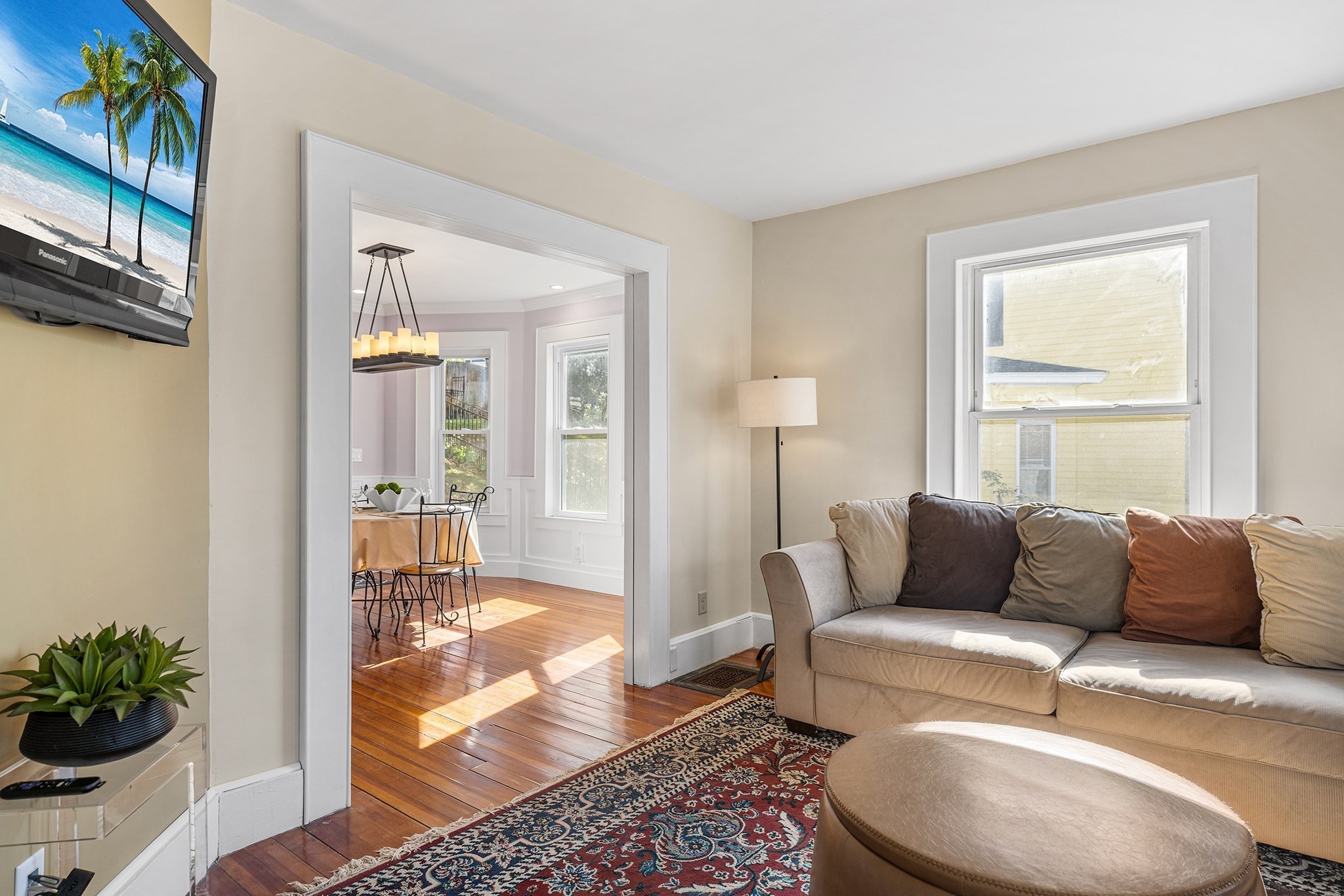 Living room with a beige sofa and patterned rug, opening into a sunlit dining room.