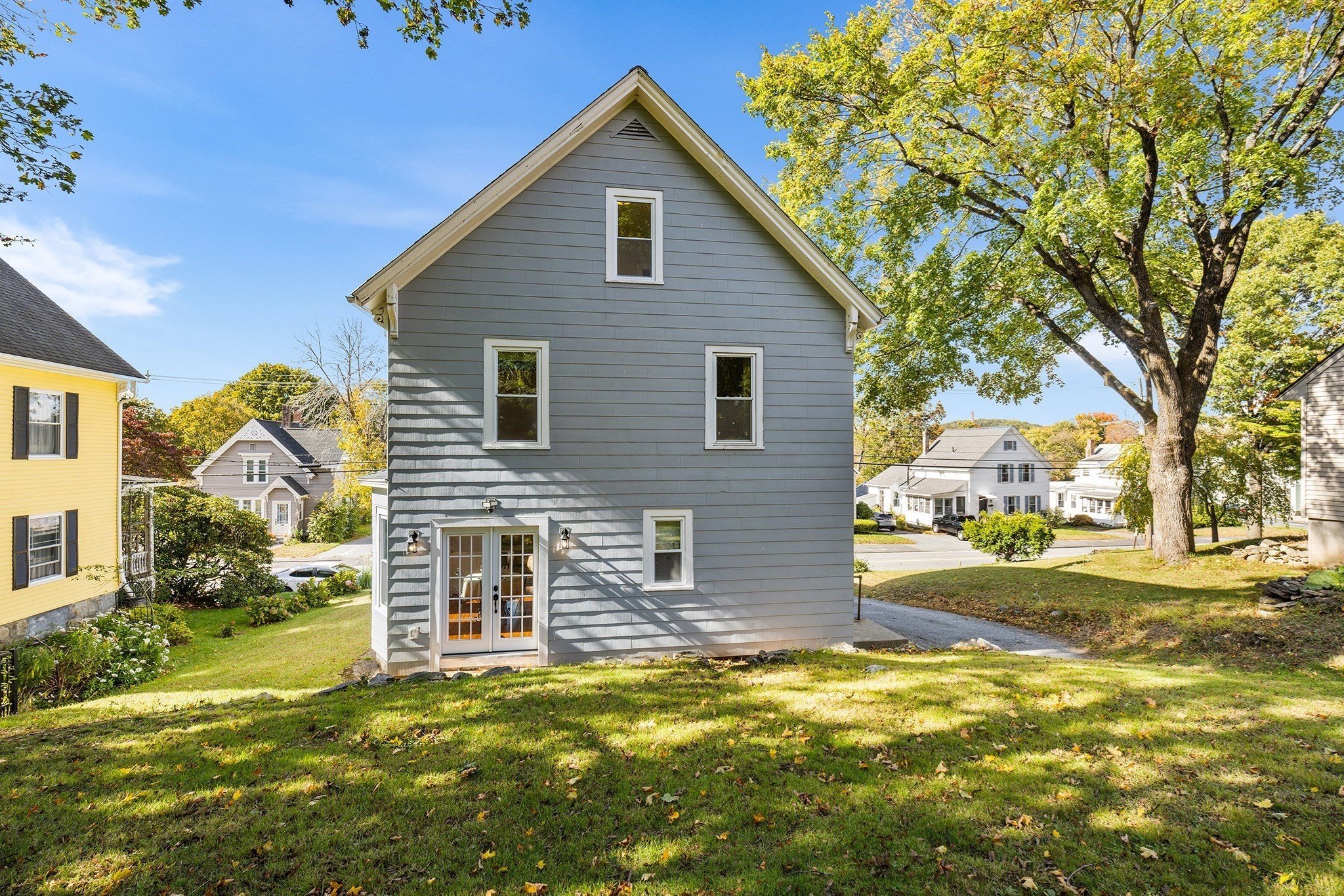 Gray two-story house with white-trimmed glass double doors, a grassy yard, and a large shade tree.
