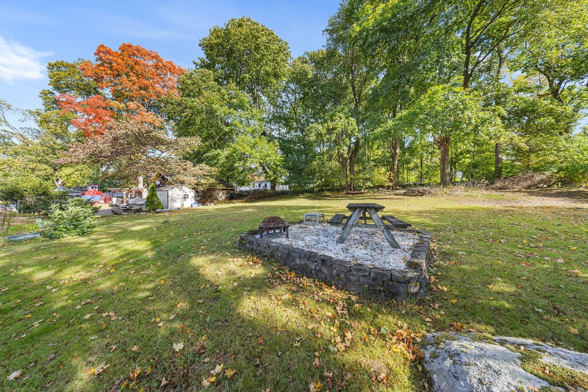Backyard with a stone-edged picnic area, wooden table, fire pit, and mature trees.
