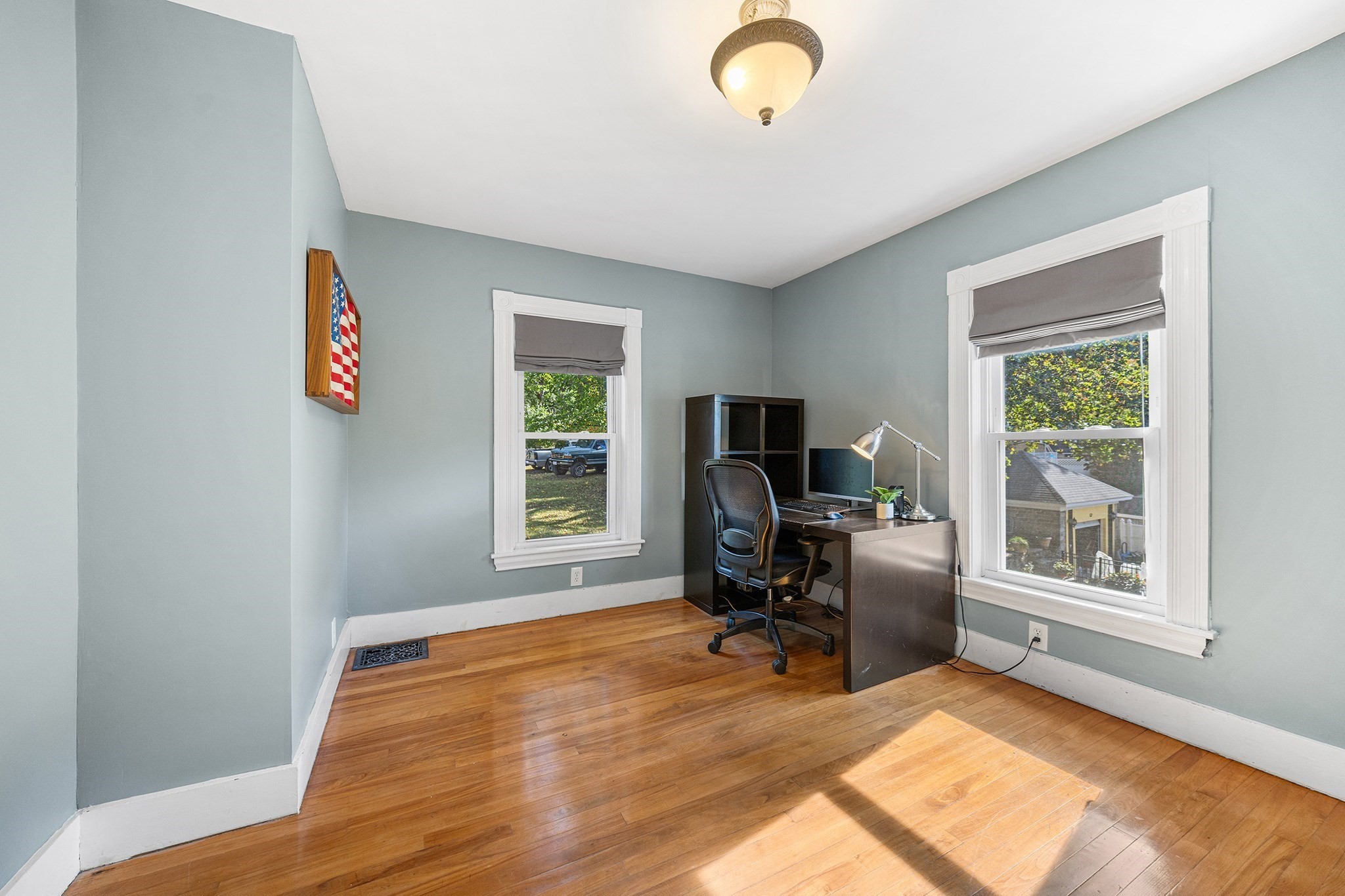 Home office with hardwood floors, two bright windows, and a desk with computer and chair.